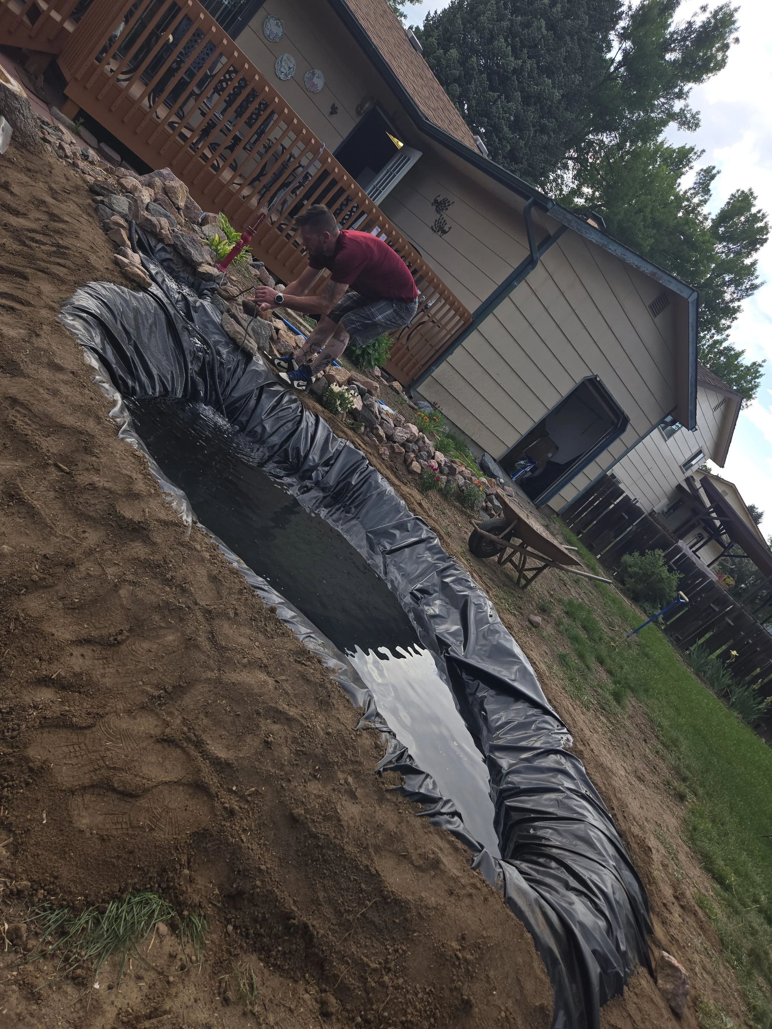 A man working on a garden water feature with a pond lined with black plastic, surrounded by rocks, in the backyard of a house with beige siding and a wooden deck.
