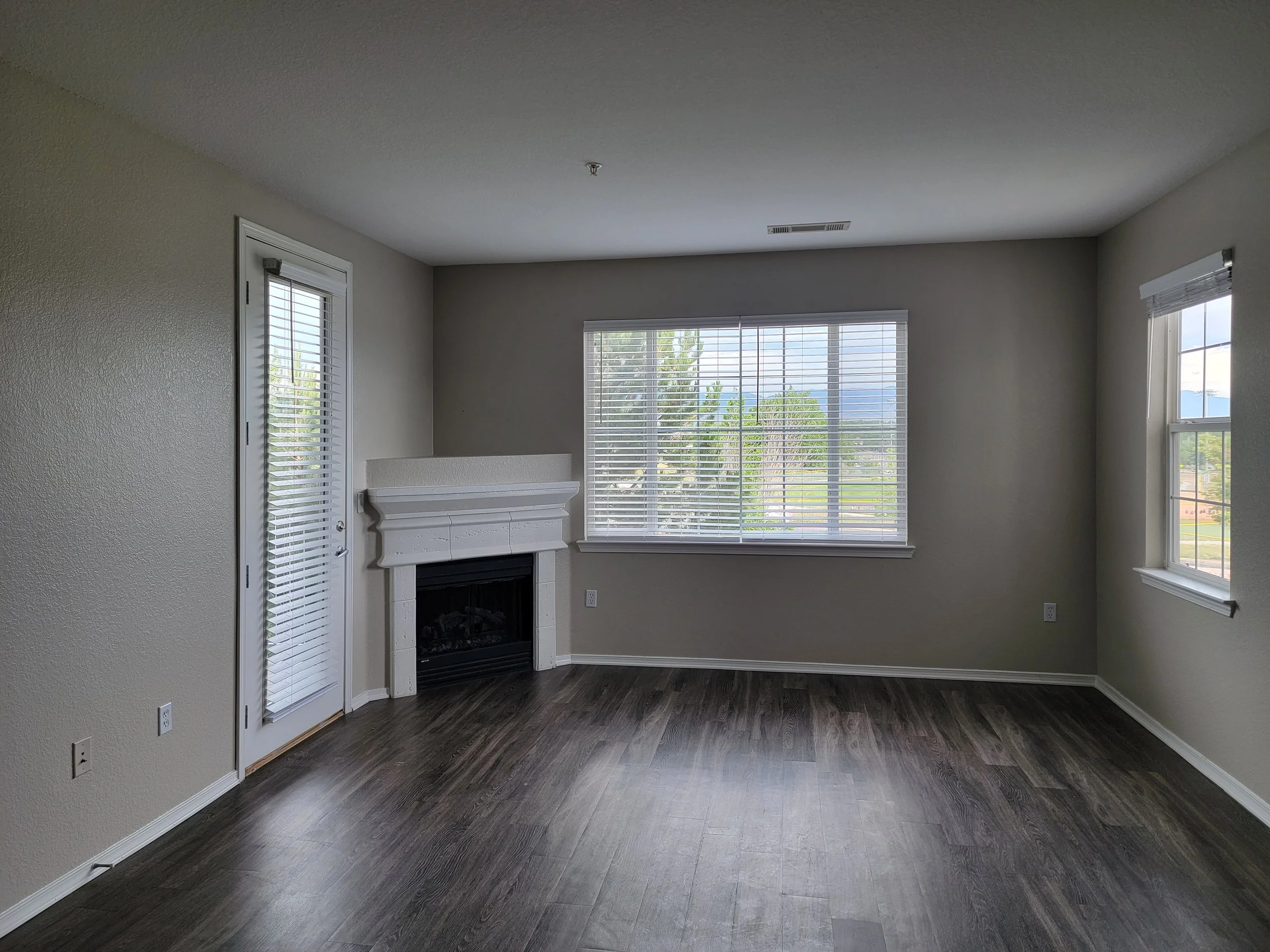 An empty room with large windows, a white fireplace, and wood flooring.
