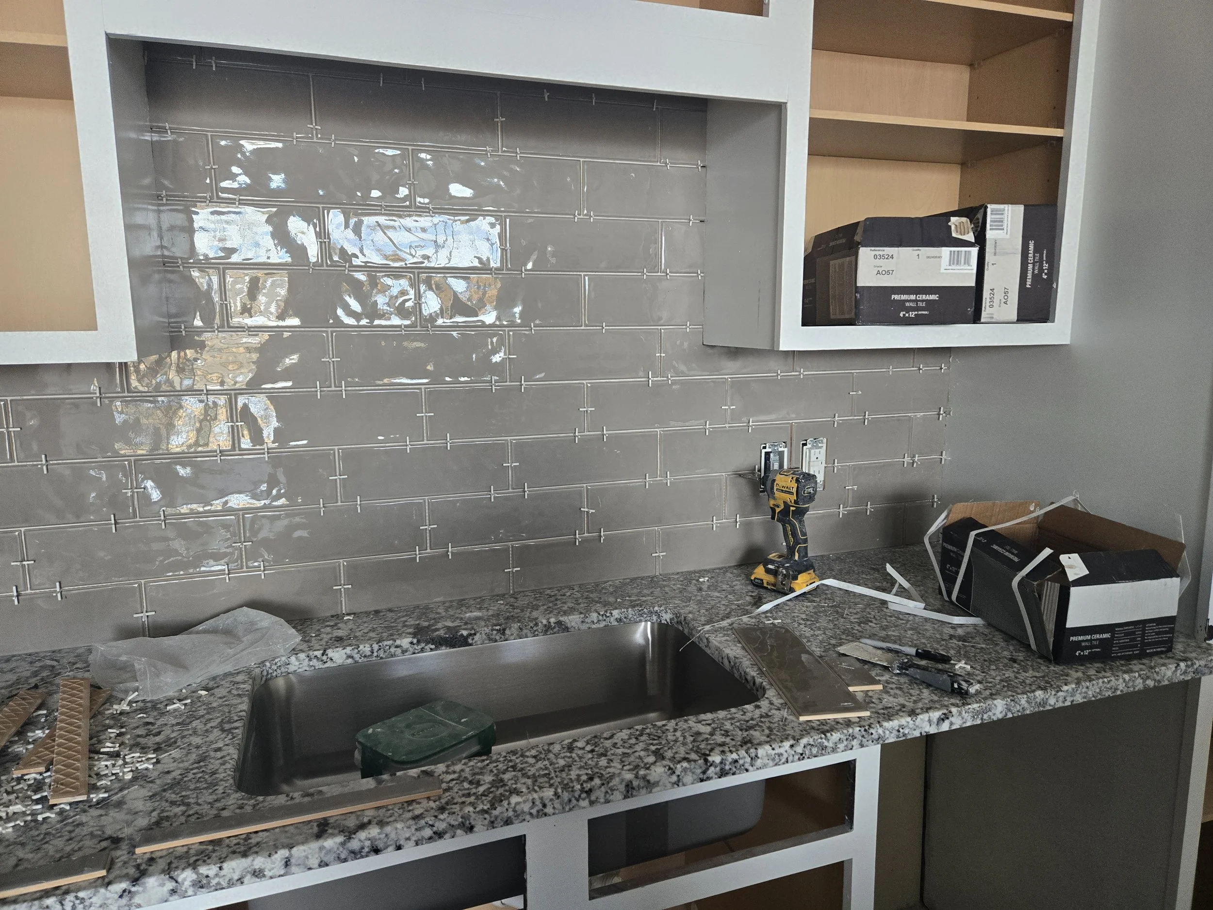 Kitchen with grey tile backsplash, granite countertop, and unfinished cabinetry. Tools and boxes are on the countertop, and the back wall has electrical outlets and wiring.