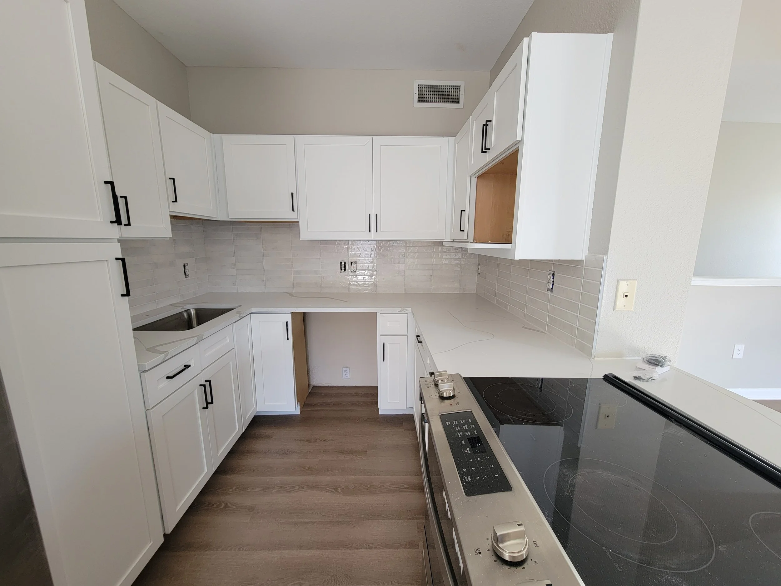 An unfinished kitchen with white cabinets, a black electric stove, a beige tile backsplash, and hardwood flooring.