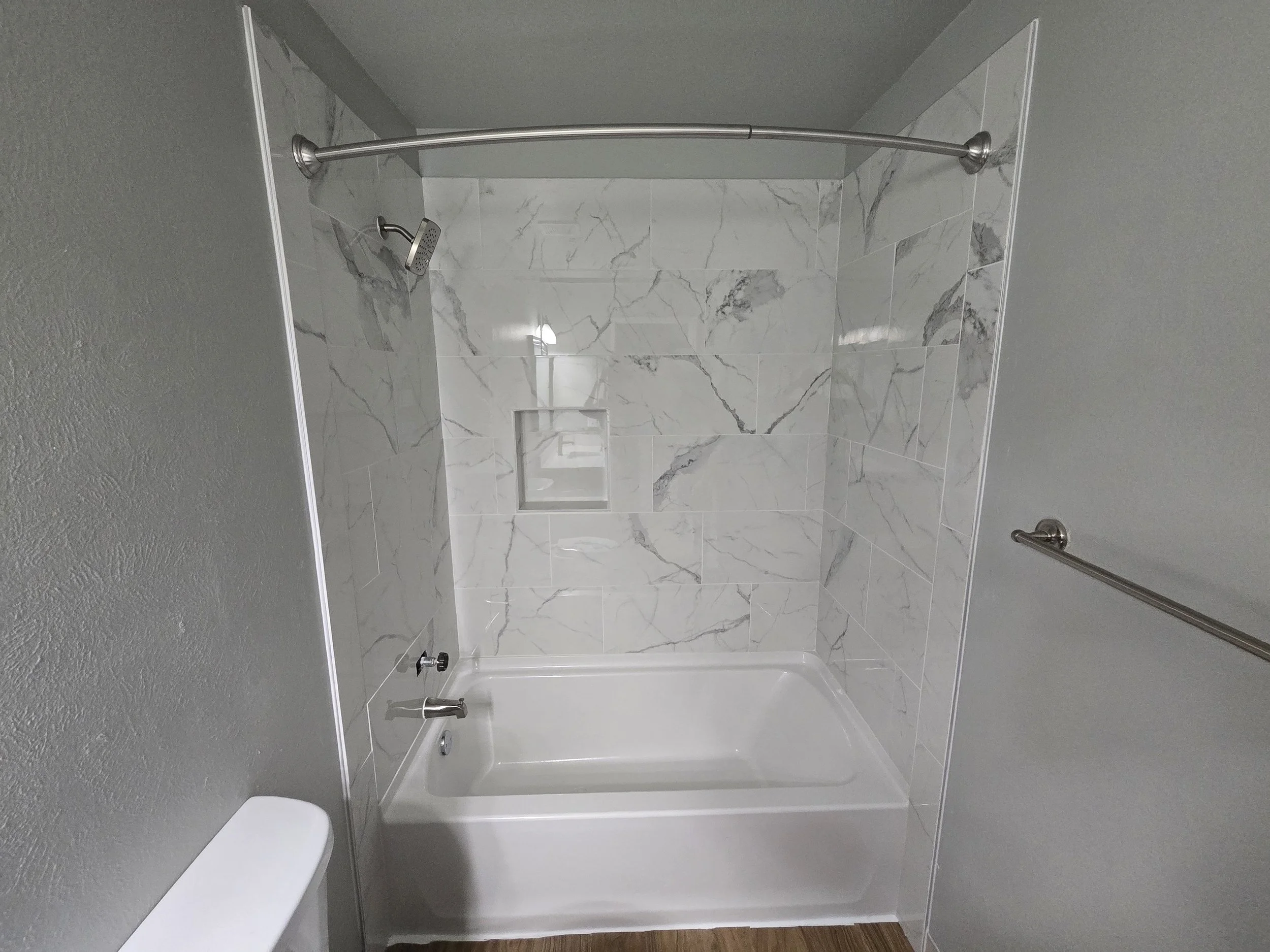 A bathroom shower area with marble tile walls, a small built-in niche, a shower head, and a curved shower curtain rod.