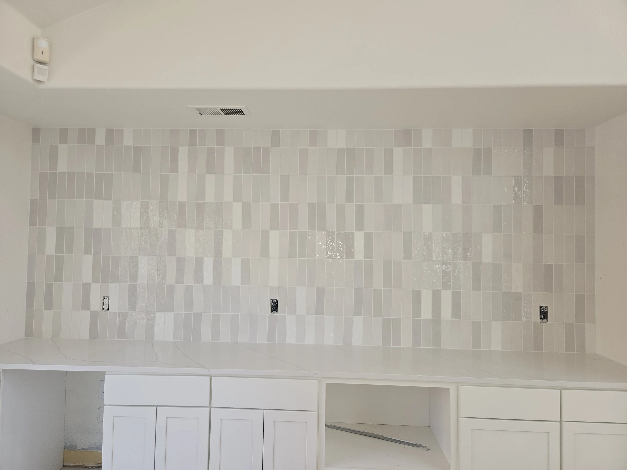 Empty kitchen space with white cabinets, beige tiled wall, and a white ceiling with vent and two electrical outlets.
