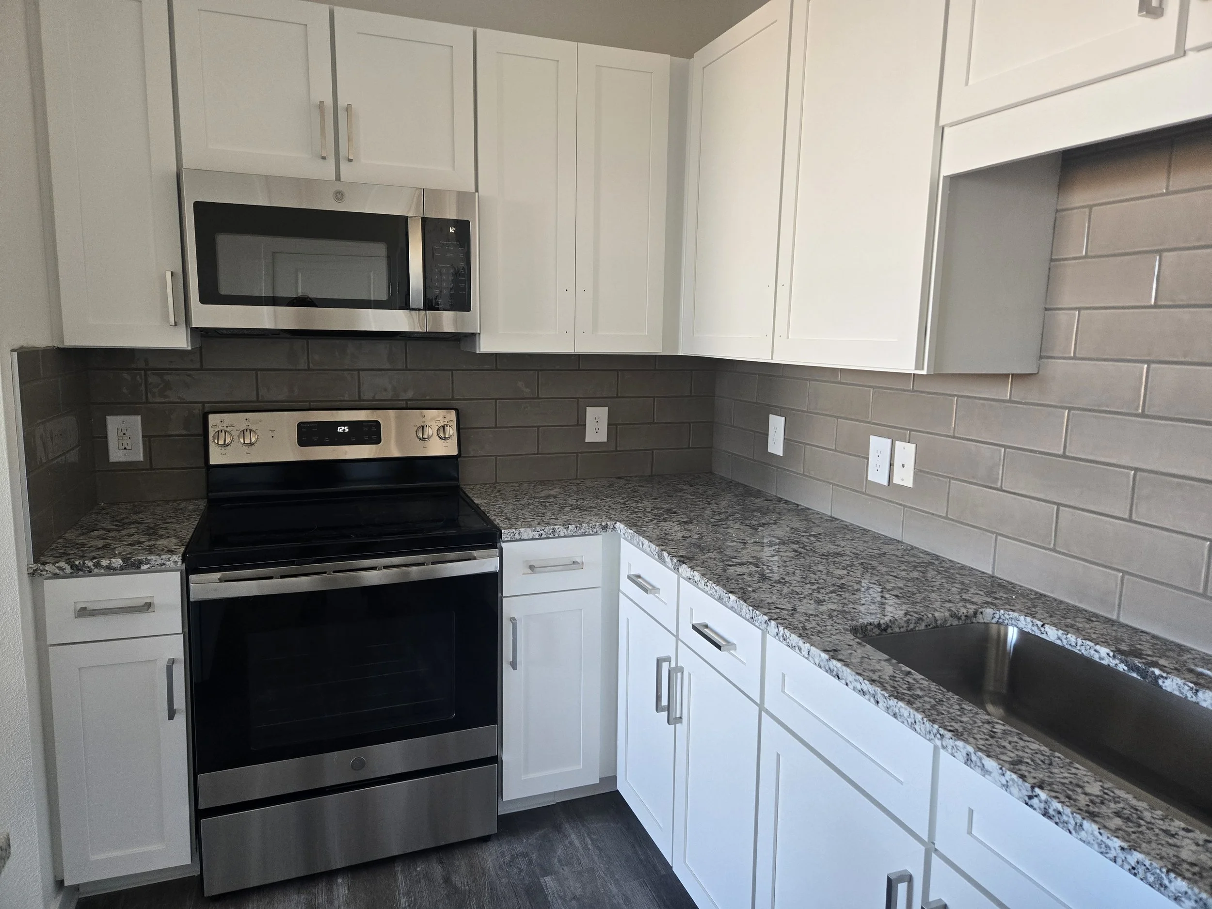 Modern kitchen with white cabinets, gray granite countertops, stainless steel appliances including a microwave and oven, gray subway tile backsplash, and multiple electrical outlets.