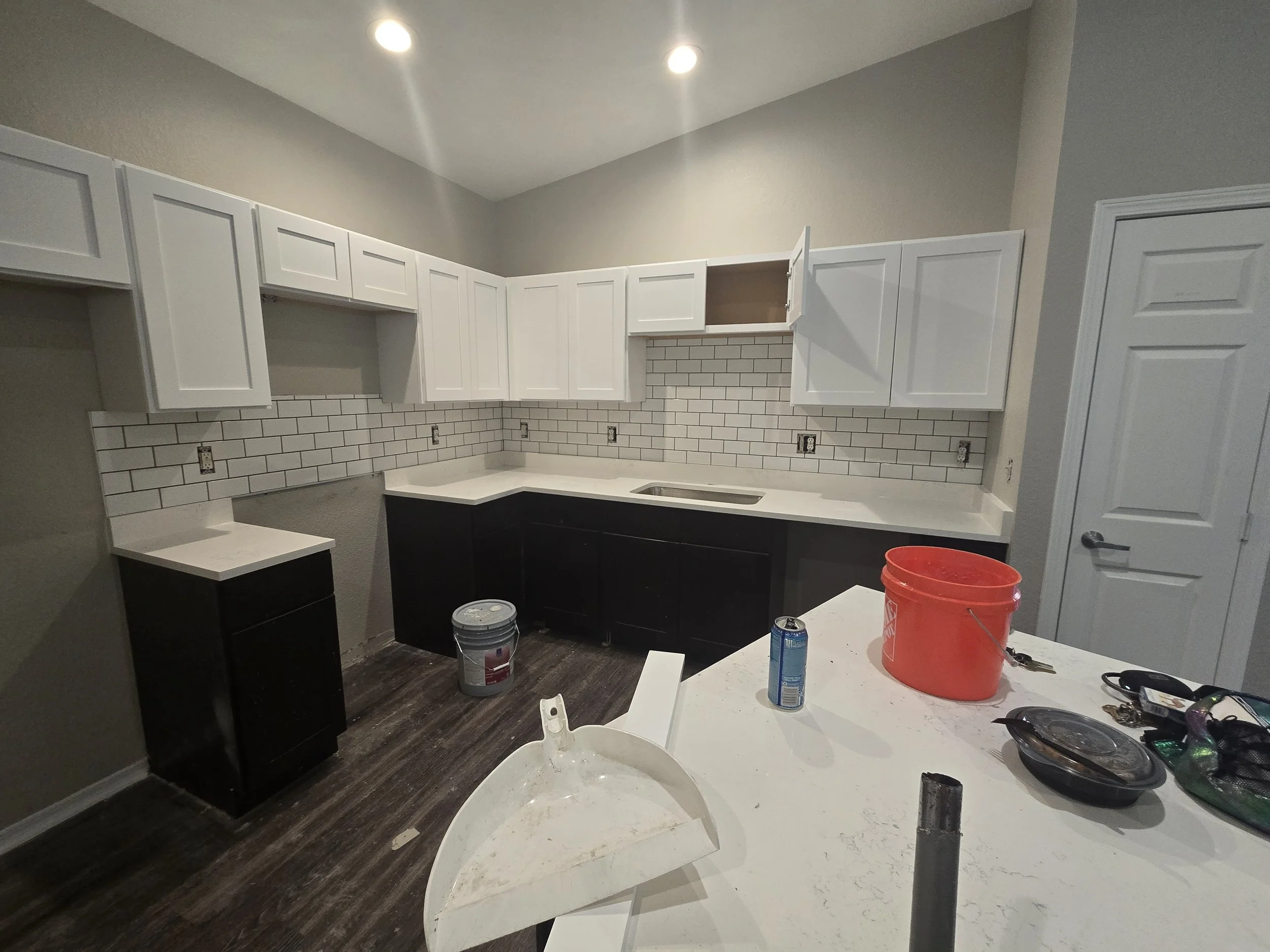 Kitchen under renovation with white upper cabinets, black lower cabinets, a white countertop, and a subway tile backsplash. Construction tools and supplies, including paint buckets, a red bucket, and various small items, are scattered on the counters
