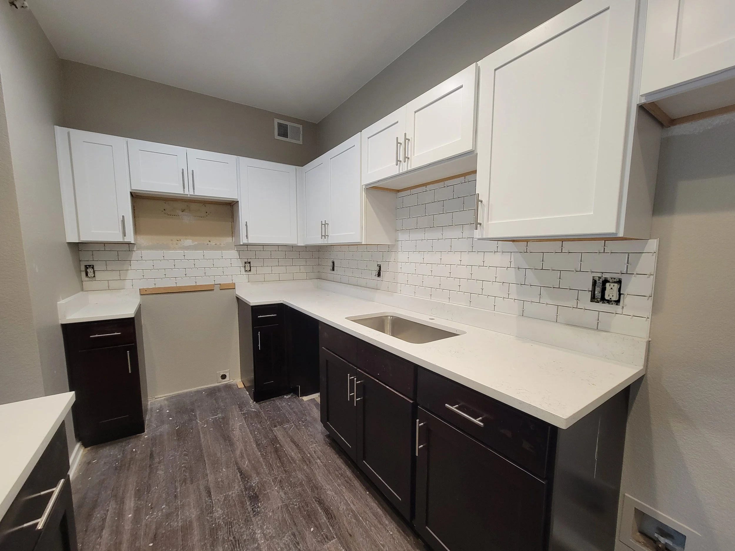 Kitchen with white upper cabinets, black lower cabinets, white countertops, a brick-patterned white tile backsplash, a sink, and electrical outlets, with some cabinets unfinished or missing doors.