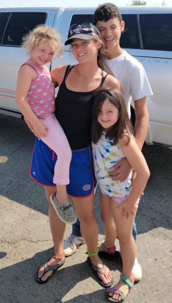 A woman and three children smiling outdoors next to a silver vehicle in sunny weather.