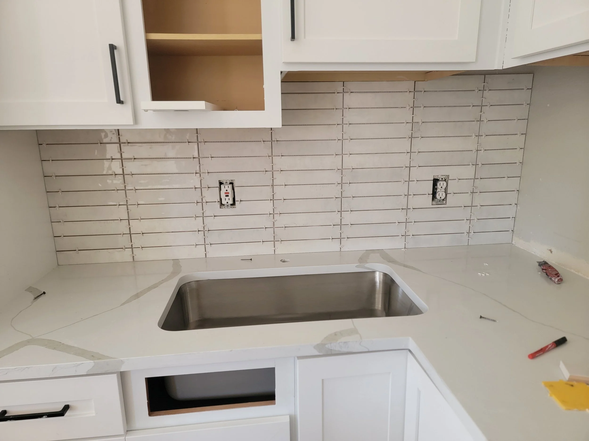 Empty kitchen cabinet with white doors, adjacent to exposed electrical outlets and a partially installed backsplash, with a kitchen sink below.