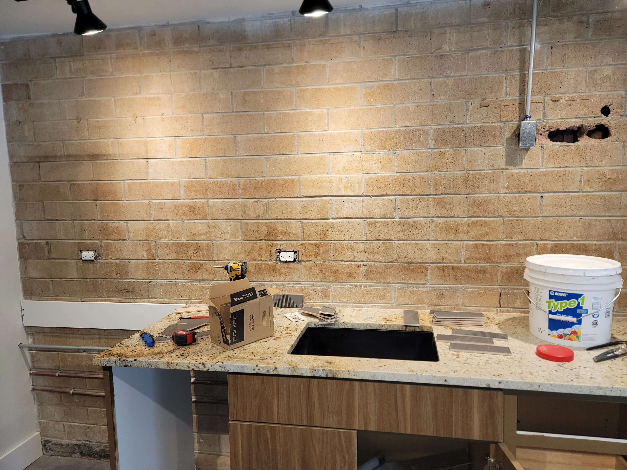 A pre renovation kitchen with a beige stone countertop, partially visible black sink, and a brick wall in the background. Construction materials are scattered on the countertop, including a yellow drill, small tiles, and a large bucket of adhesive.
