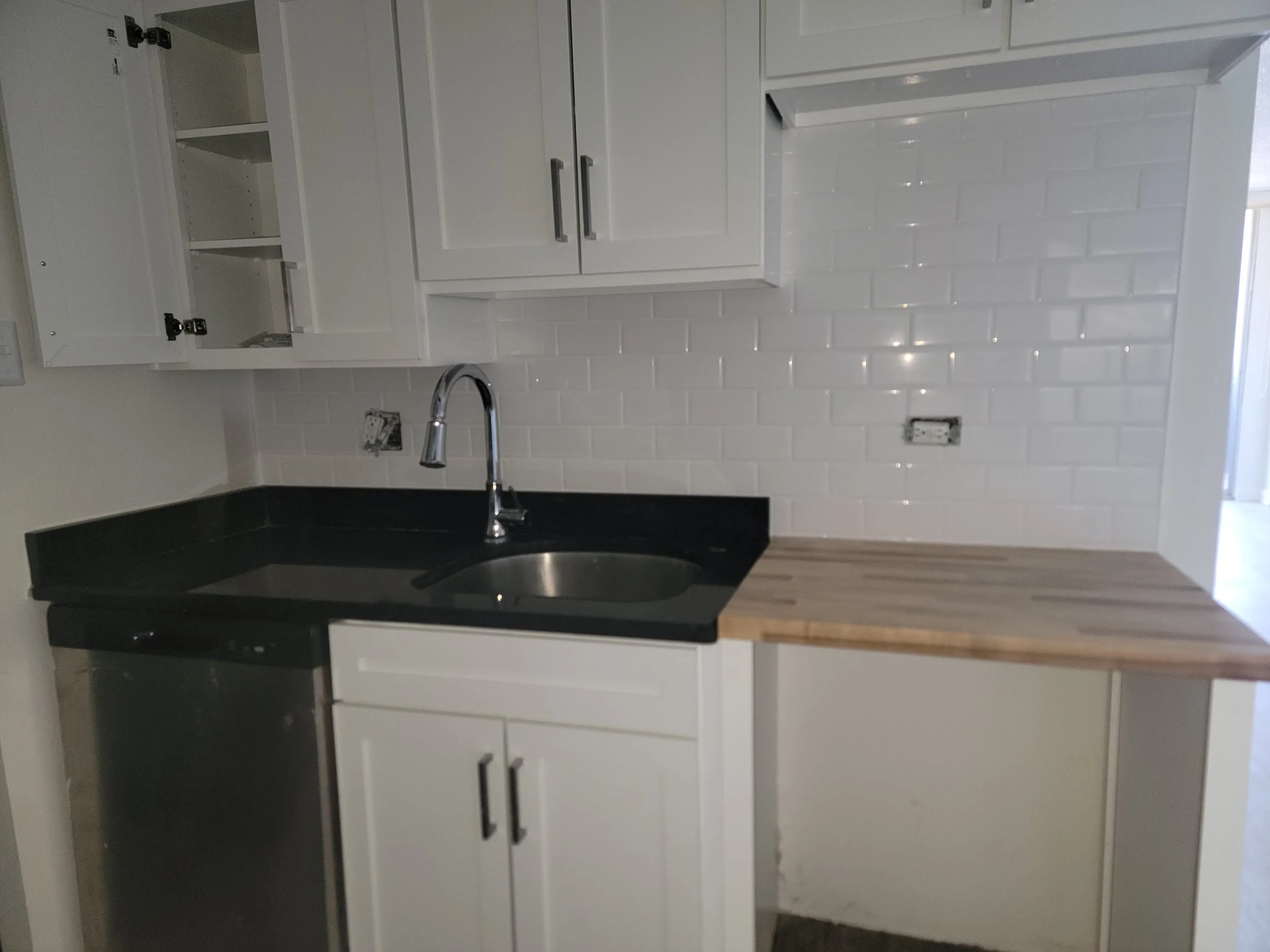 A partly finished kitchen with white cabinets, a black countertop, a double sink with a chrome faucet, and a white subway tile backsplash. The upper cabinets are open, and there is an electrical outlet on the wall.