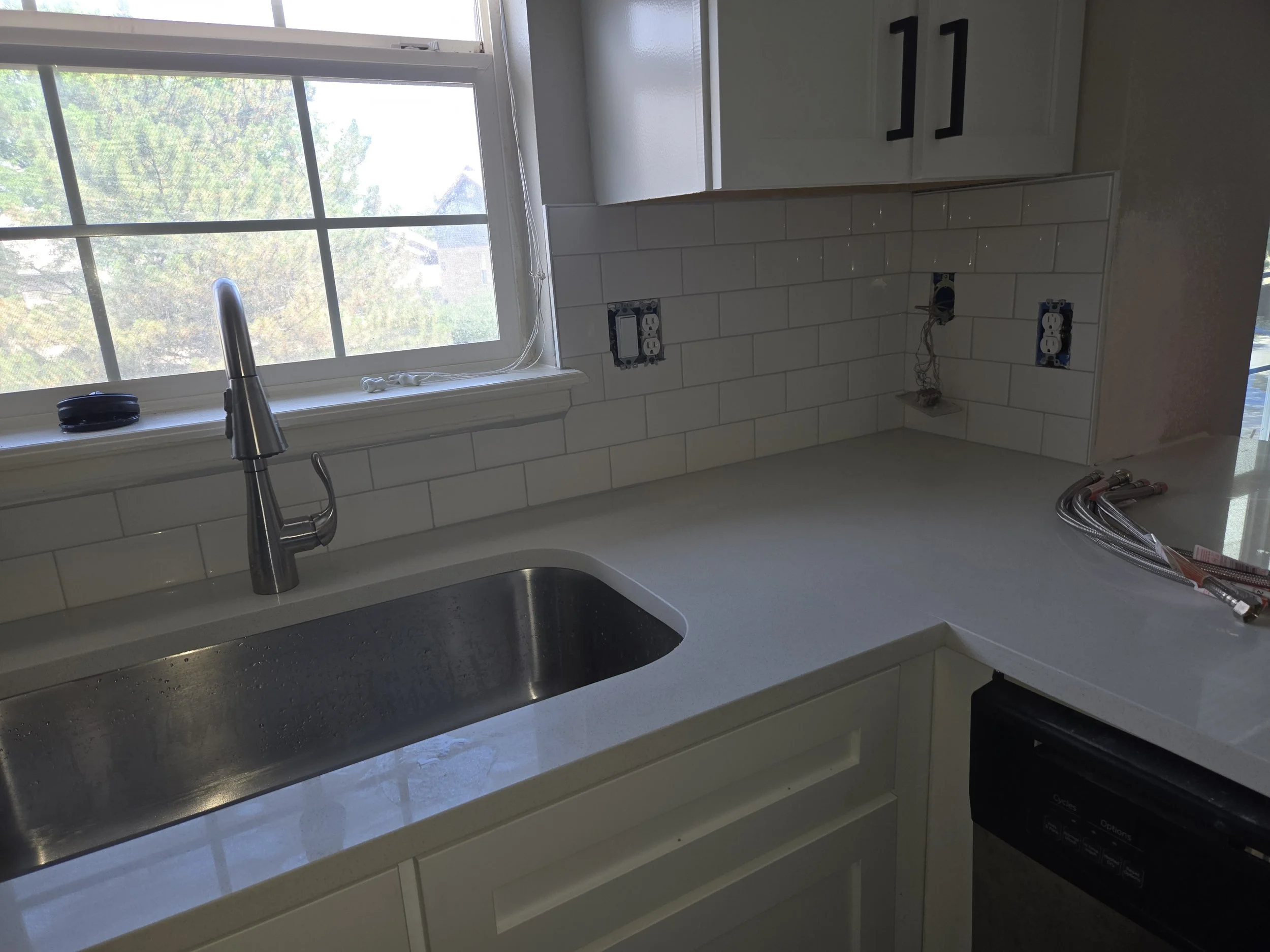 Kitchen countertop with a stainless steel sink, a faucet, and an empty space for a stove or appliance. There are white cabinets and a window overlooking trees outside. Electrical outlets and wires are visible on the wall.