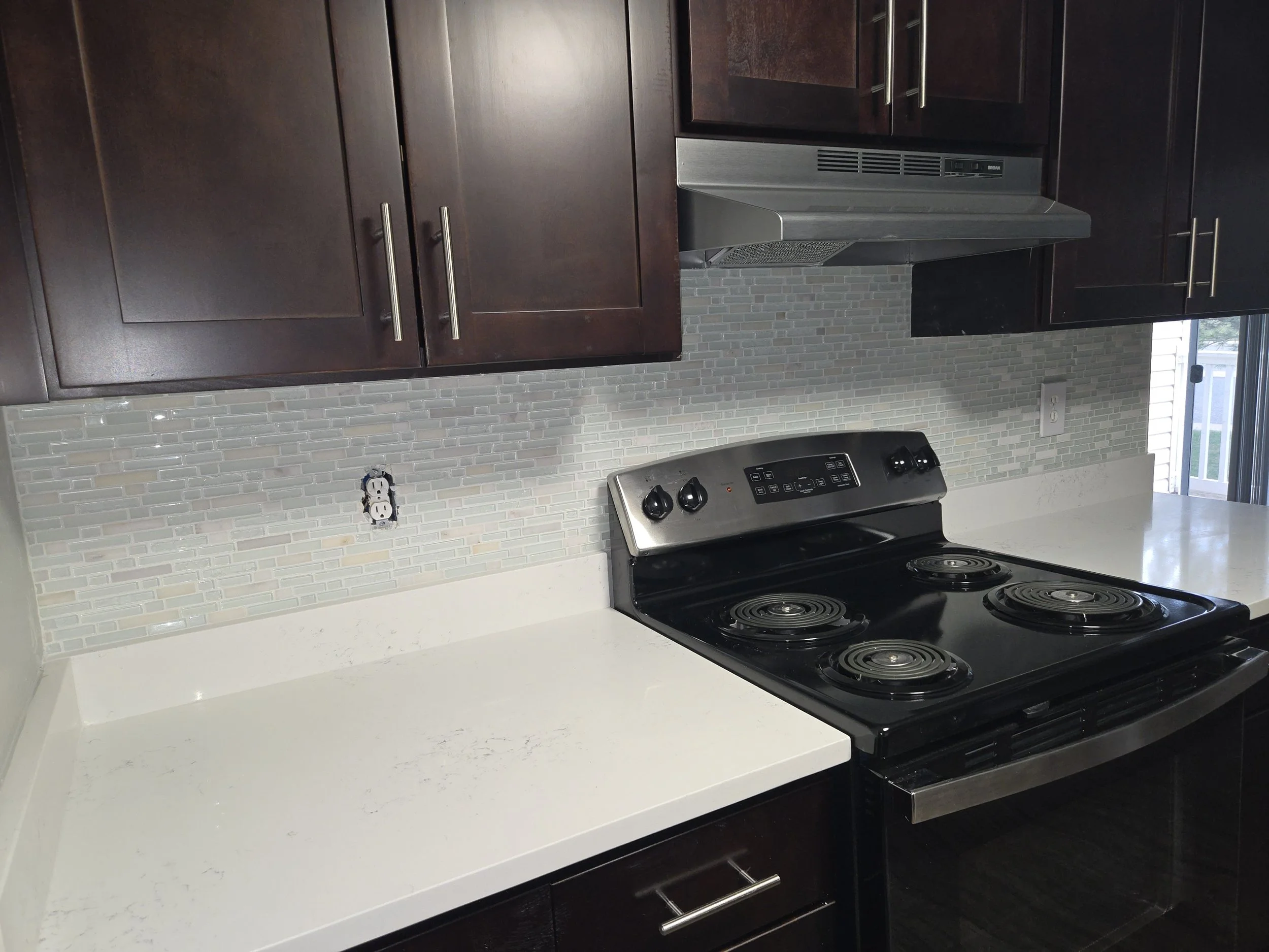 Kitchen with dark wood cabinets, stainless steel range hood, black stove with four coils, and a white countertop. The backsplash has light-colored small rectangular tiles, and part of a window is visible on the right.