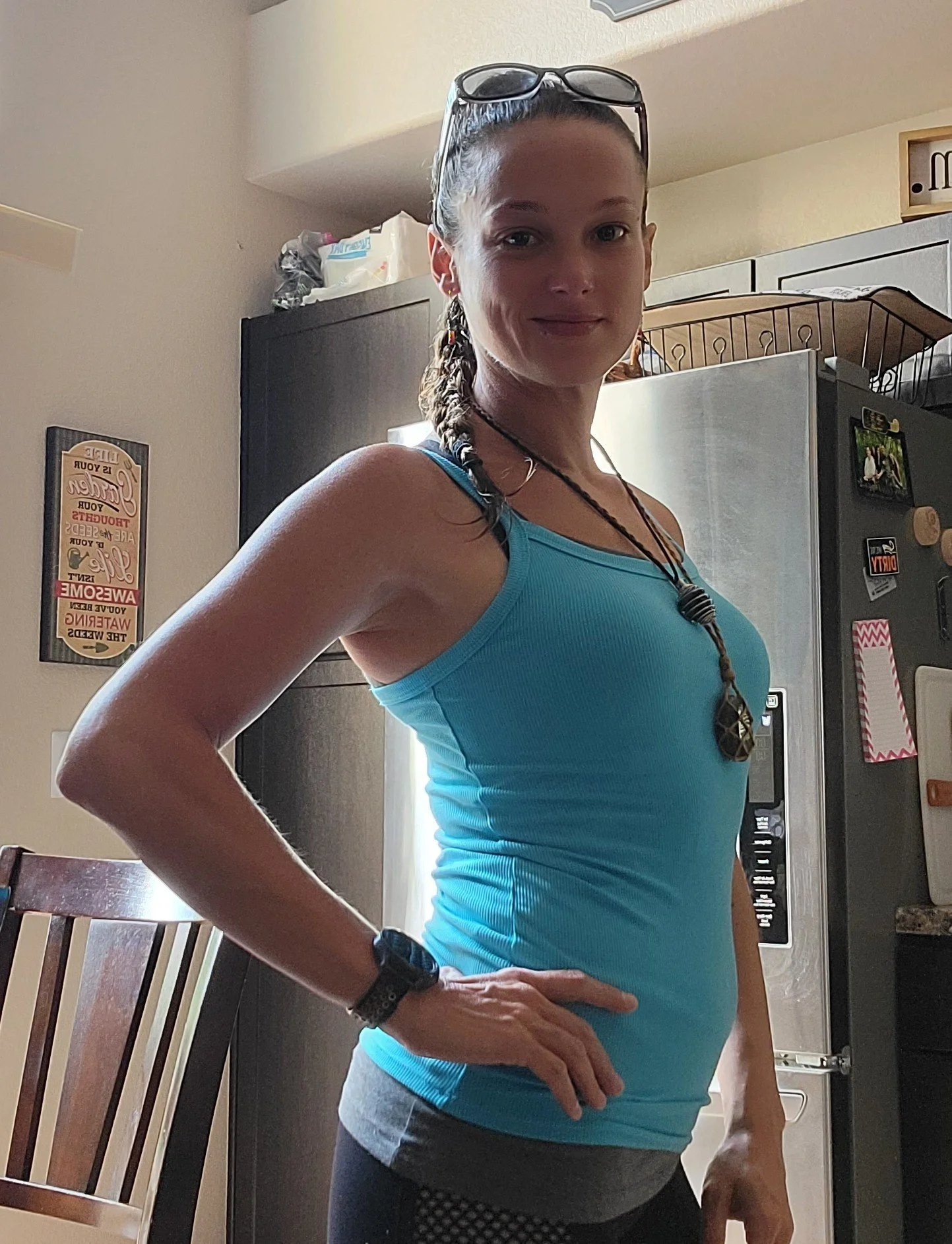 A woman wearing a blue athletic tank top, glasses on her head, bracelets, and a necklace, standing in a kitchen with her hand on her hip.