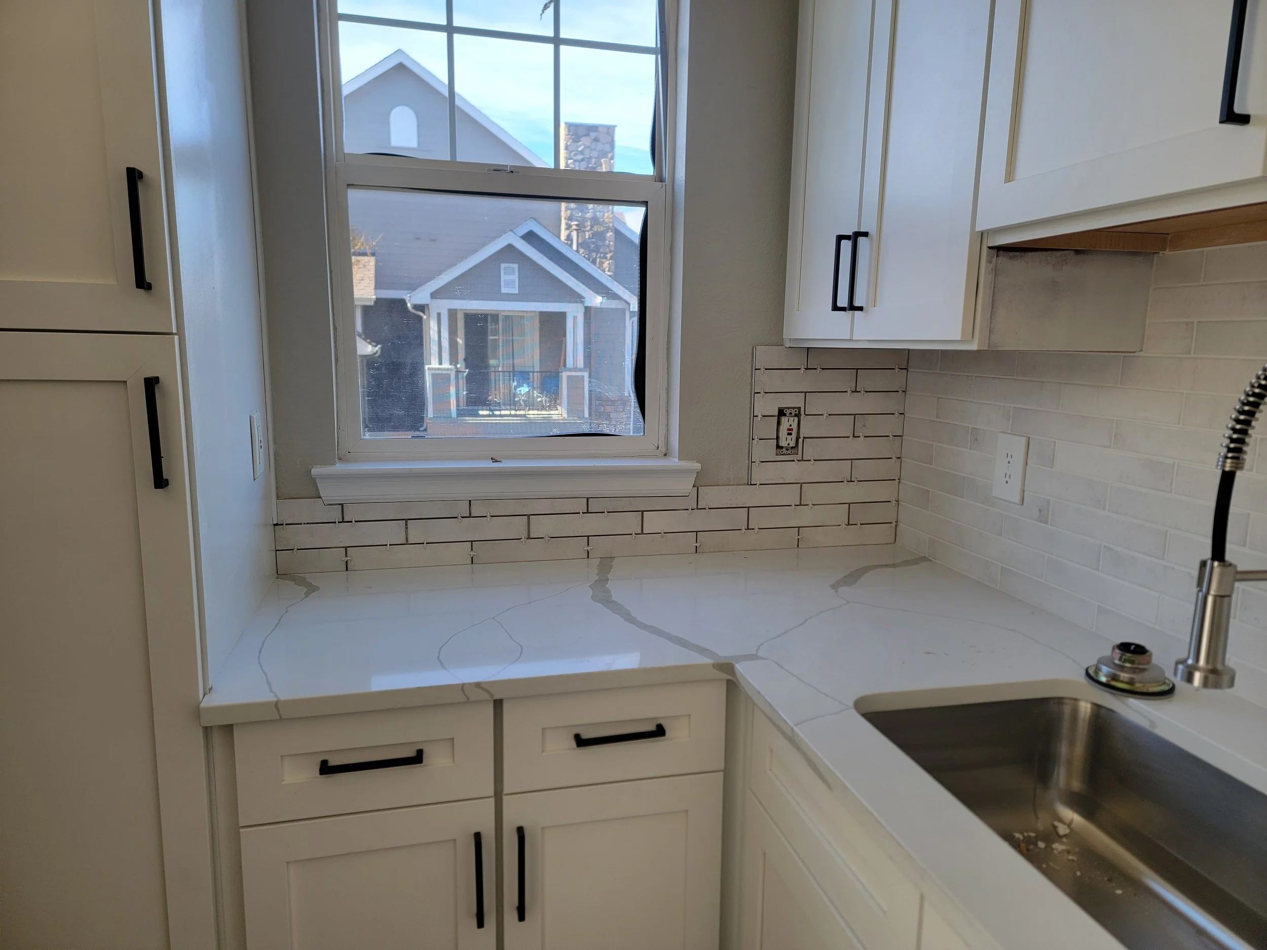 A kitchen with white cabinets, a window showing neighboring houses, a white tile backsplash, and a kitchen sink with a faucet. The countertop is white, and there is an electrical outlet on the wall.