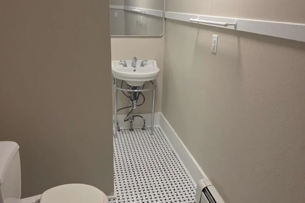 Small bathroom with a white sink, mirror, light beige walls, a grab bar, and a toilet partially visible in the foreground.