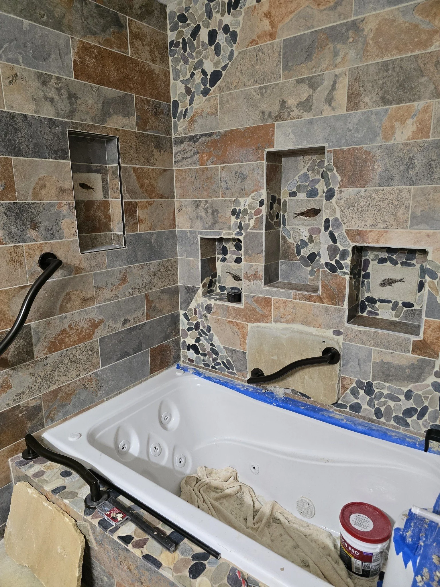 An unfinished bathtub surrounded by multi-colored tile walls with inlaid pebble accents. Tools and supplies are scattered inside and around the tub, including a towel and a container of joint compound.