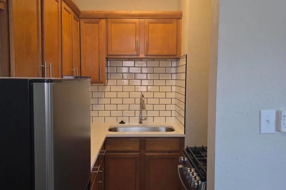 Small kitchen corner with wooden cabinets, a white countertop, a stainless steel sink, white subway tile backsplash, a black stove, and a partially visible black refrigerator.