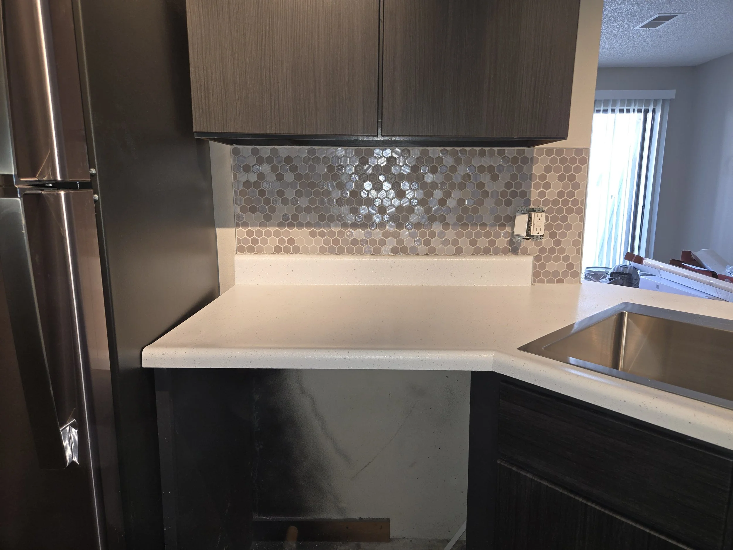 Kitchen countertop with a backsplash of small hexagonal tiles in shades of gray, a white lower countertop, dark wooden cabinets above, a silver refrigerator on the left, and a small electrical outlet on the wall. Part of a sliding glass door with ver