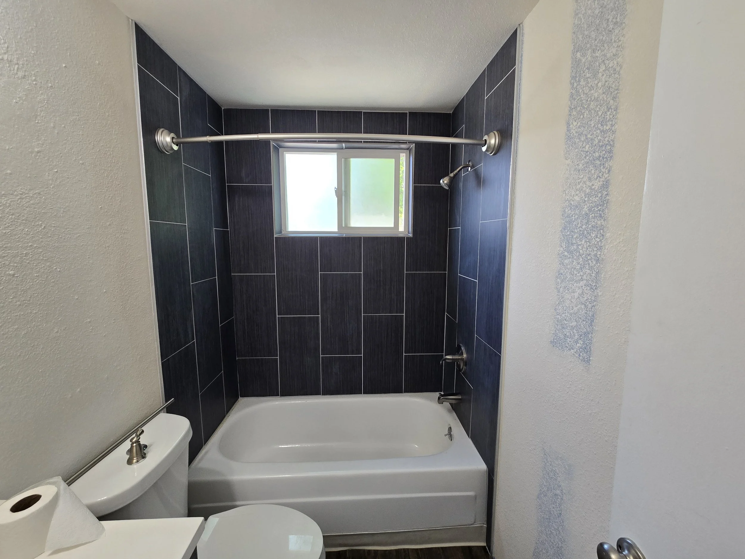Bathroom with black tiled walls, a small window, a bathtub, a shower head, a toilet with a toilet paper roll, and a towel bar without any towels.