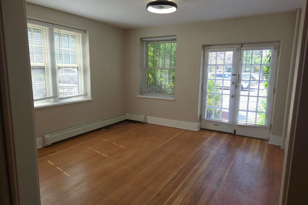 Empty living room with hardwood floors, three large windows with blinds, a glass door with blinds, and a ceiling light fixture.