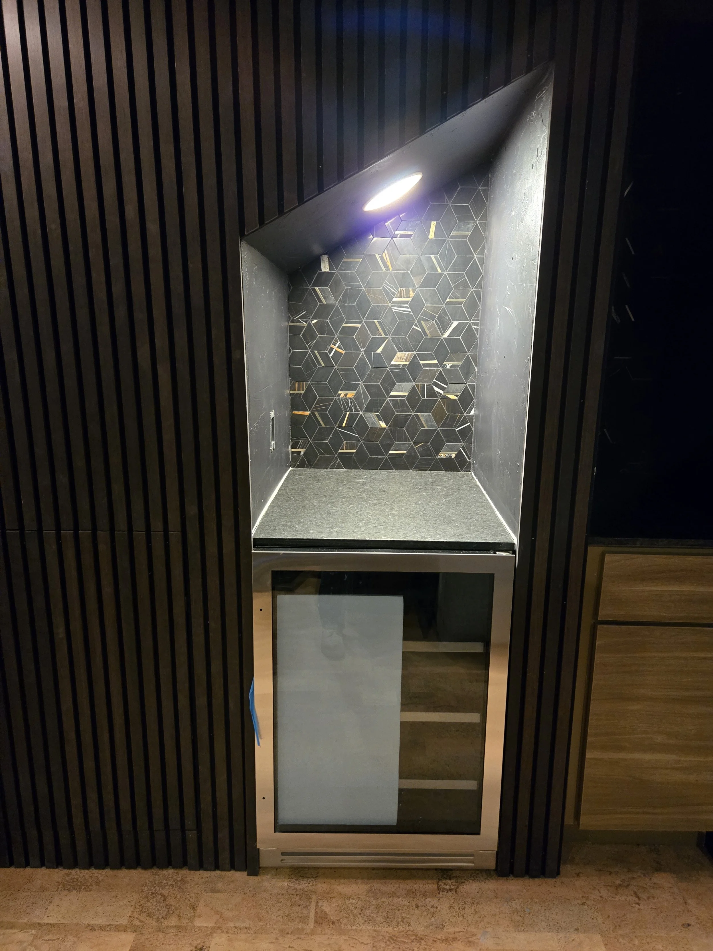 Empty wet bar with black interior walls featuring vertical stripes and a backsplash with black and gold hexagonal mosaic tiles, illuminated by a ceiling light.