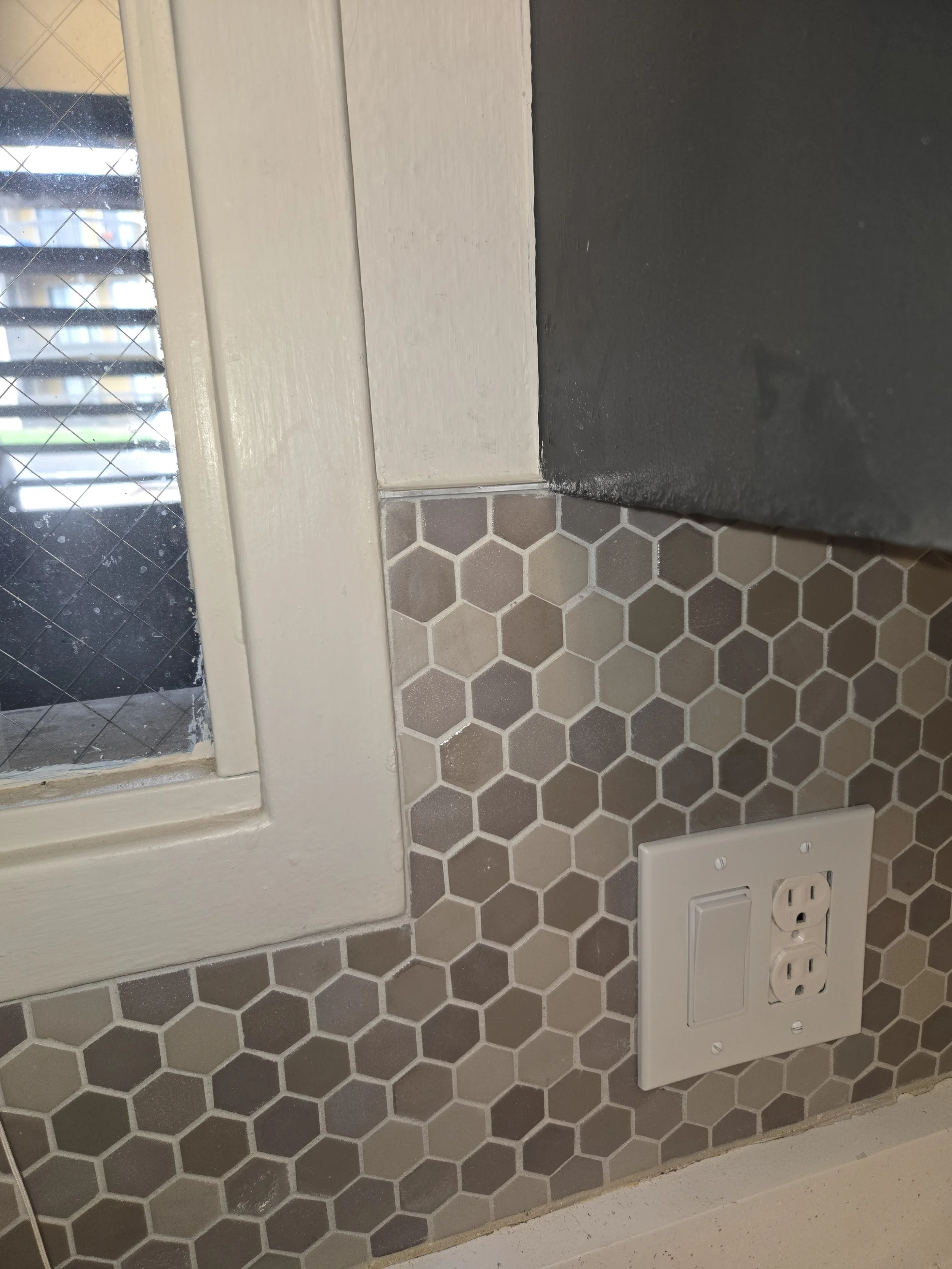 Close-up of a window with a black and dark gray painted sill, hexagonal gray and beige tiles on the wall, and a white electrical outlet with a cover plate.