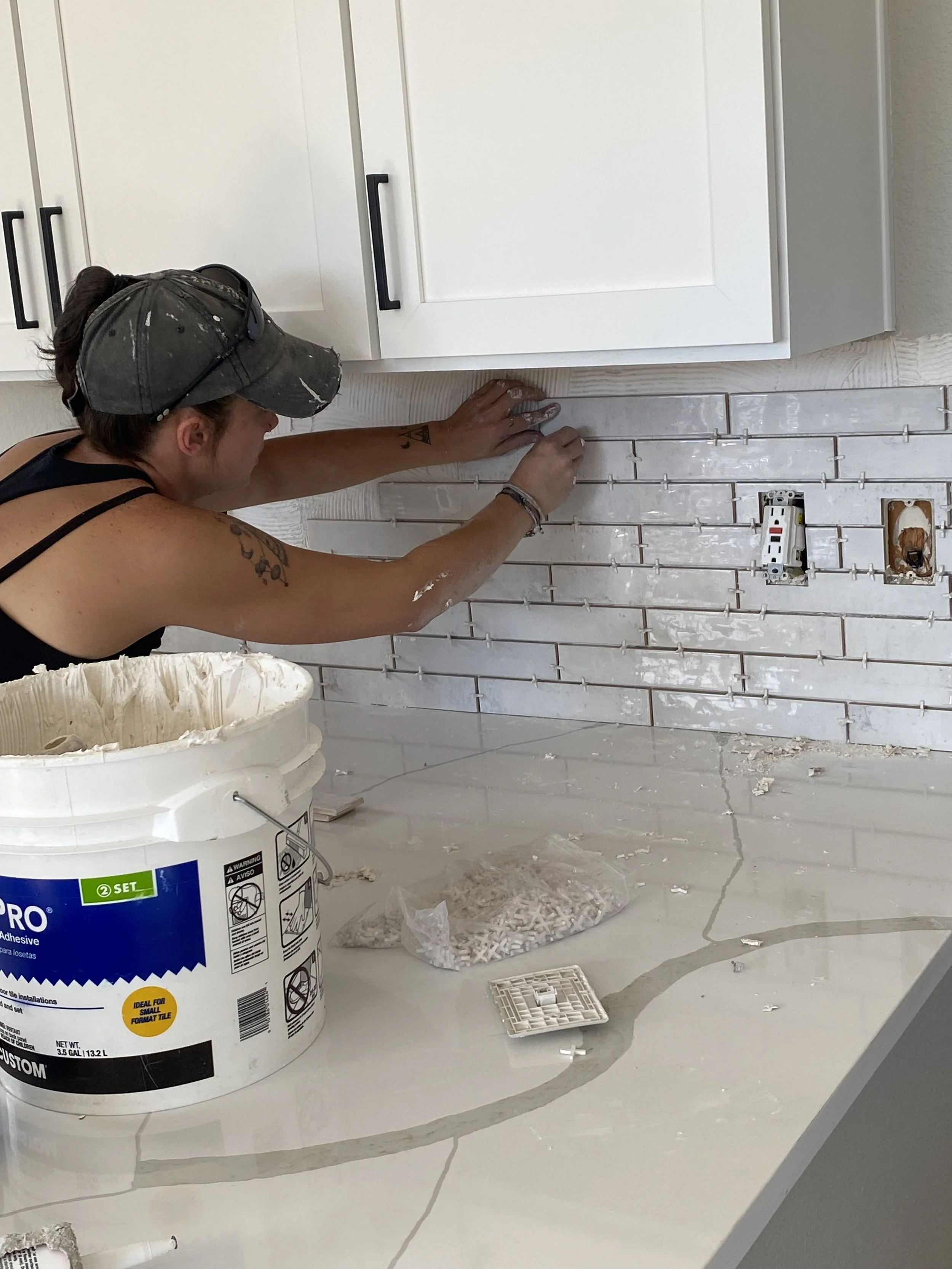 A person installing white rectangular subway tiles on a wall in a kitchen or bathroom. The person is wearing a gray cap and a black tank top, with tattoos on their arm. There is a large bucket of adhesive, and some tile spacers and tools on the countertop.