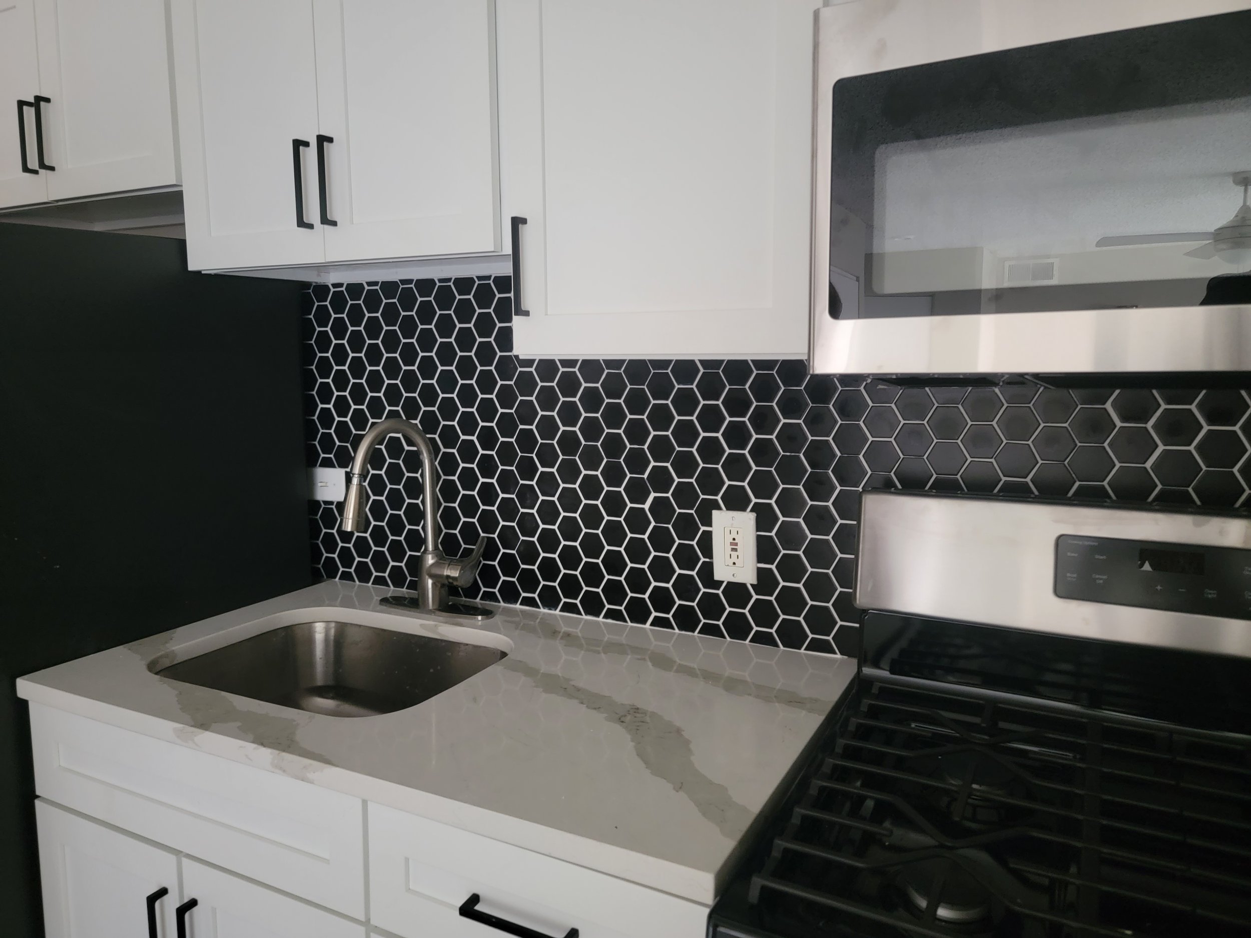 Modern kitchen with white cabinets, black hardware, black hexagonal tile backsplash, white countertop, stainless steel sink with silver faucet, black gas stove, and white electrical outlet.