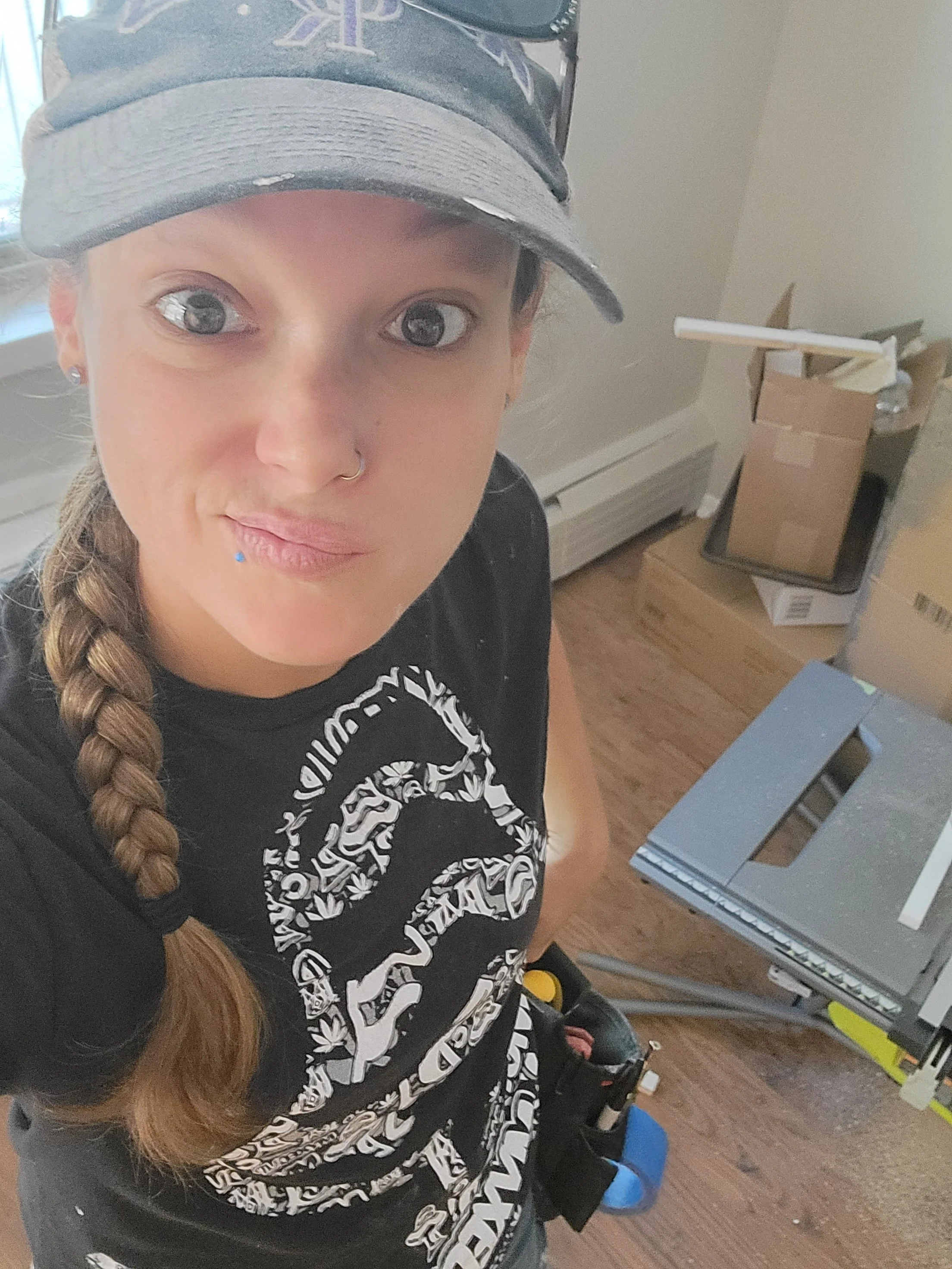 A person with long braided hair wearing a cap and a black T-shirt sitting in a room with a box, a small table, and some items on it. They are looking at the camera, and part of their face and clothing are visible.