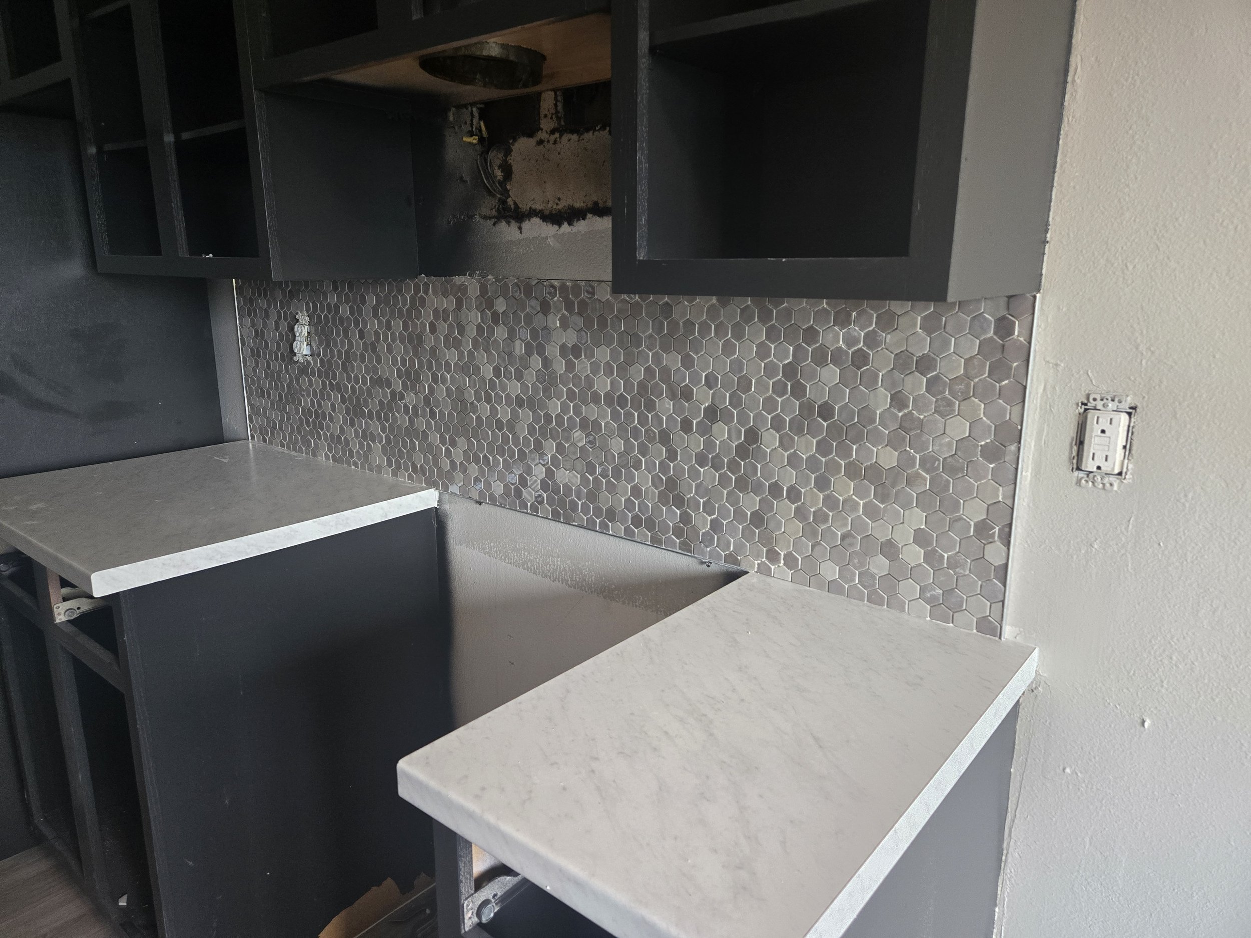 A kitchen cabinet with the door removed, revealing the interior which is stained and dirty. The surrounding area has a hexagonal tile backsplash and a wall outlet.