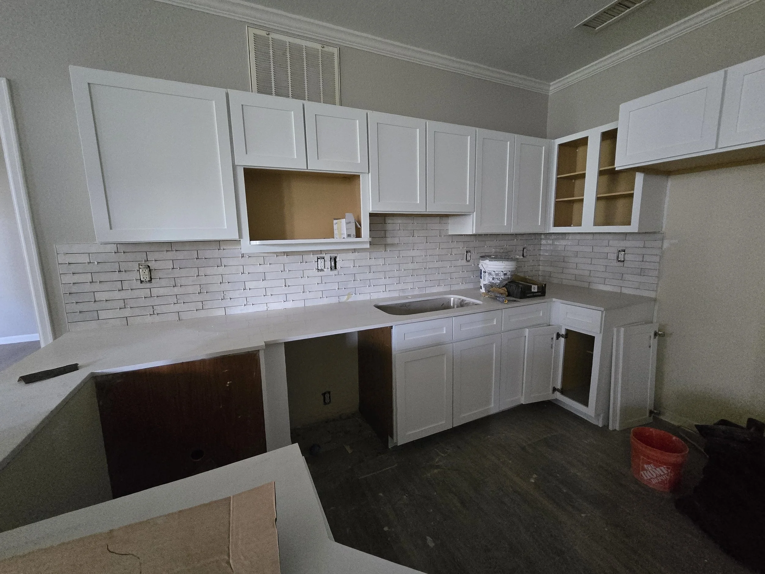 Kitchen underway with white cabinetry and a brick backsplash, with some cabinets missing doors, construction tools, and an orange bucket on the dark hardwood floor.
