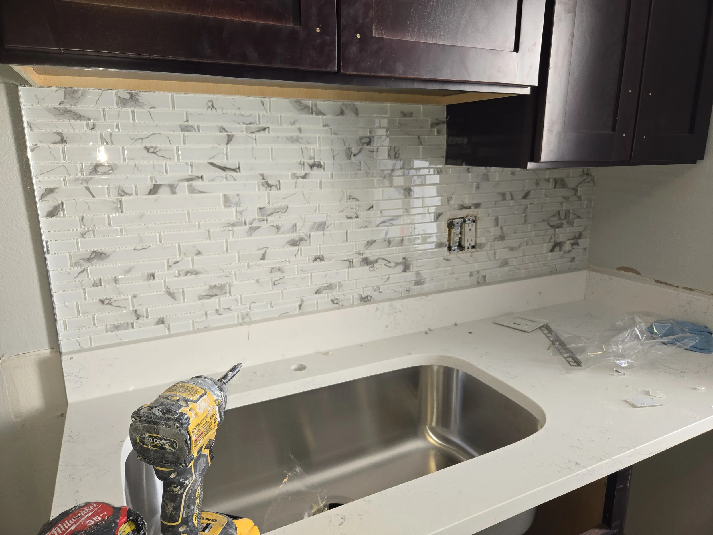 Kitchen sink area with a marble backsplash, dark upper cabinets, a white countertop, and a cordless drill placed on the counter.