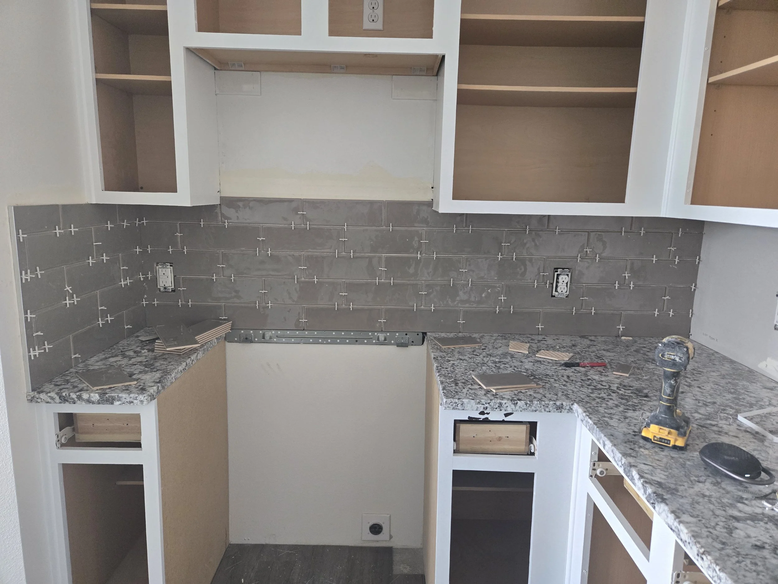 Kitchen cabinets under construction with white framing, some shelves and drawers installed, and tools like a drill on the granite countertop.