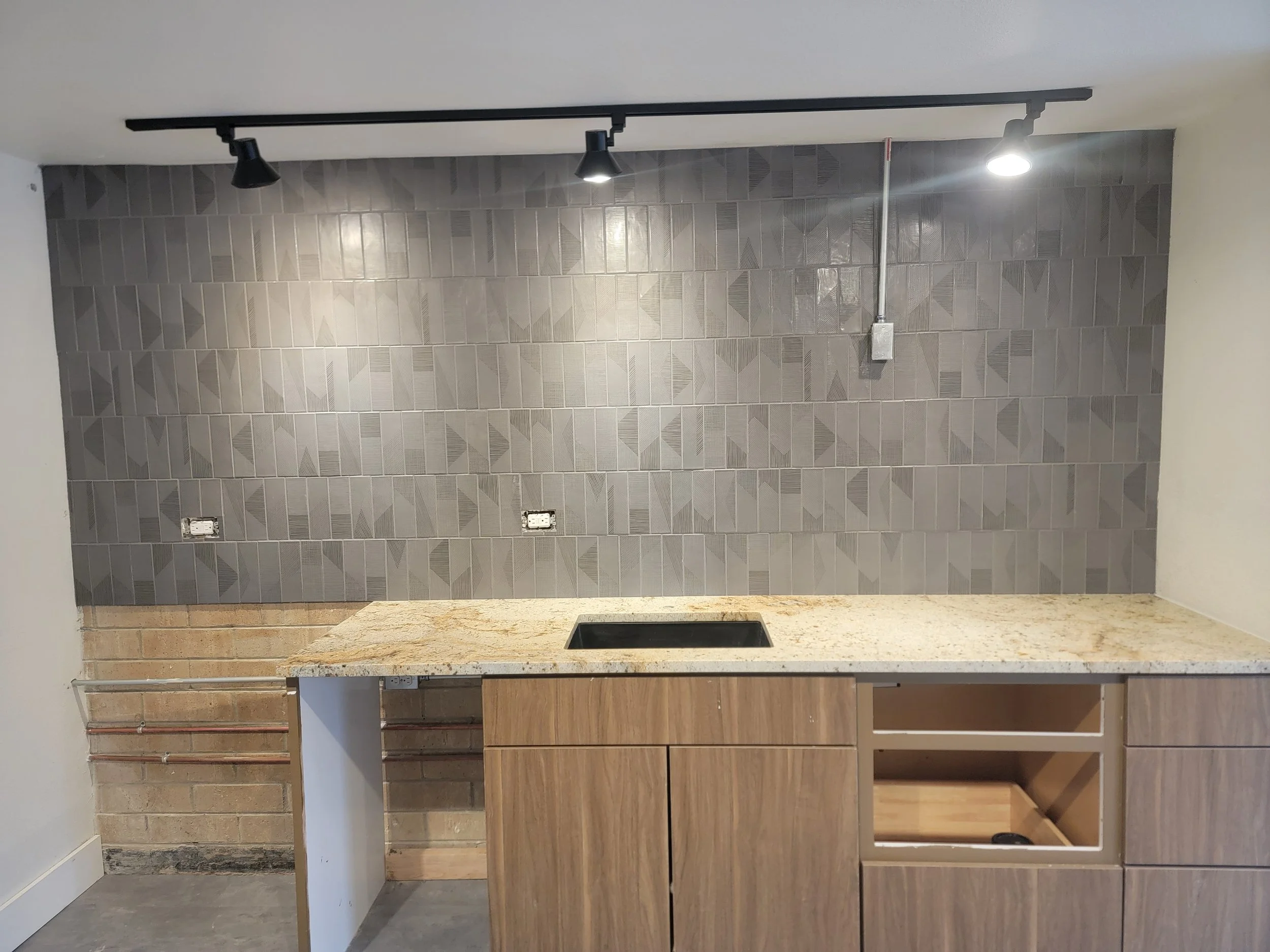 A post renovation kitchen with a beige granite island, wood cabinets, a gray tiled wall, and ceiling track lighting.