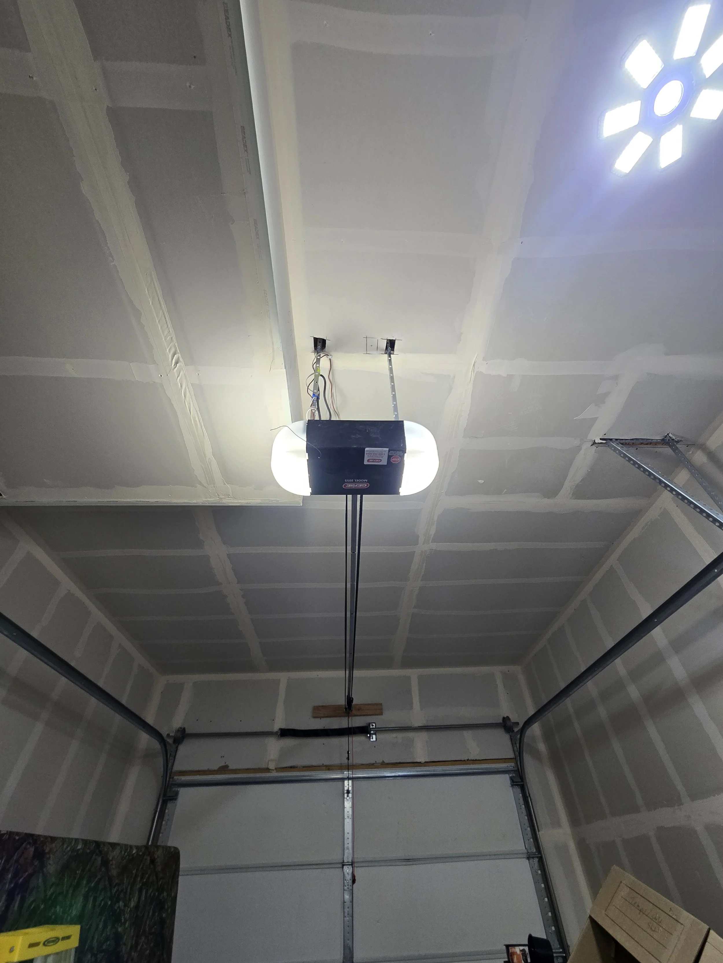 The interior of a garage with drywall walls and ceiling, an overhead garage door opener with exposed wiring, and a window with decorative cutouts on the ceiling