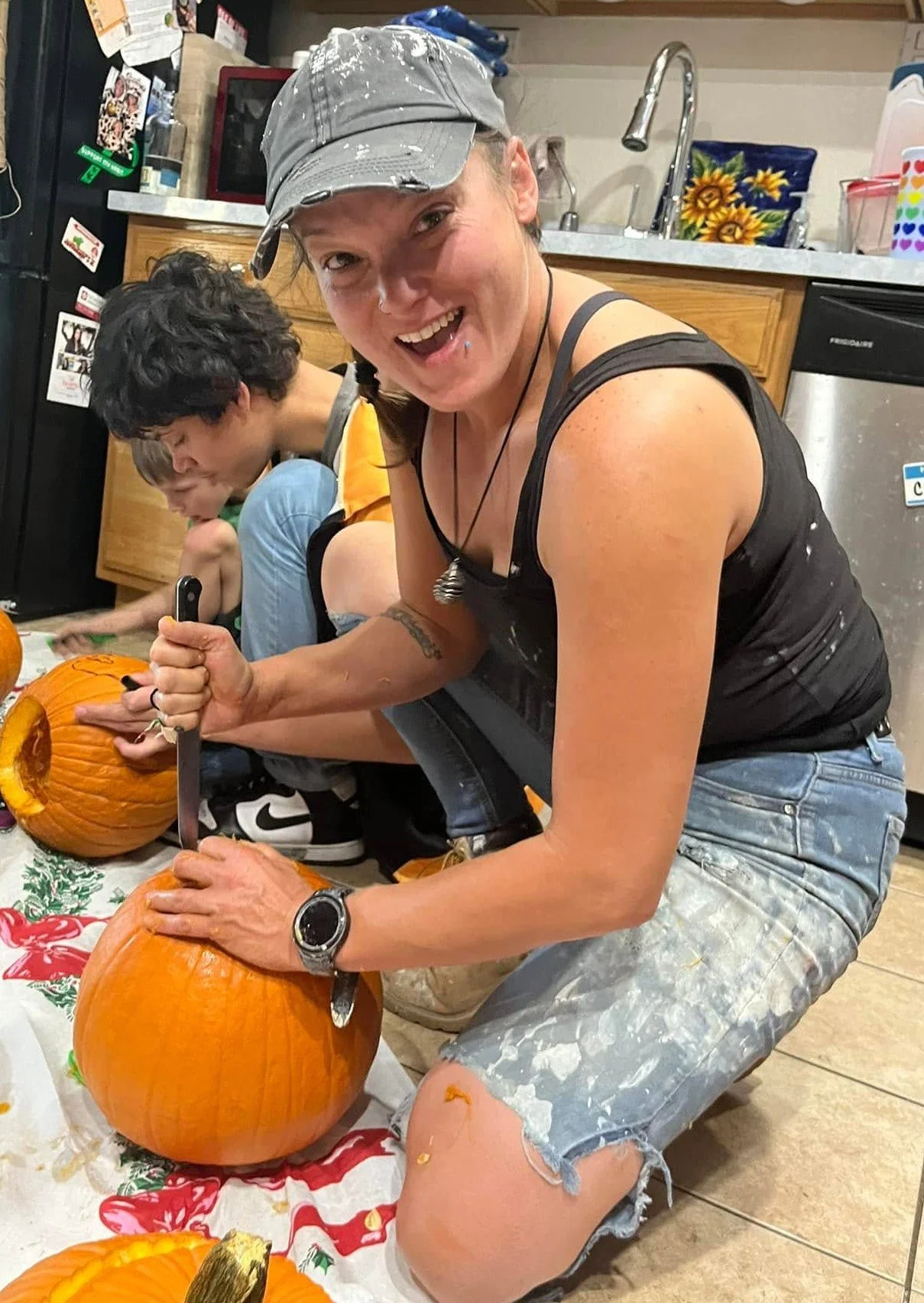 A woman smiling and carving pumpkins at a kitchen table, with two children in the background also carving pumpkins.