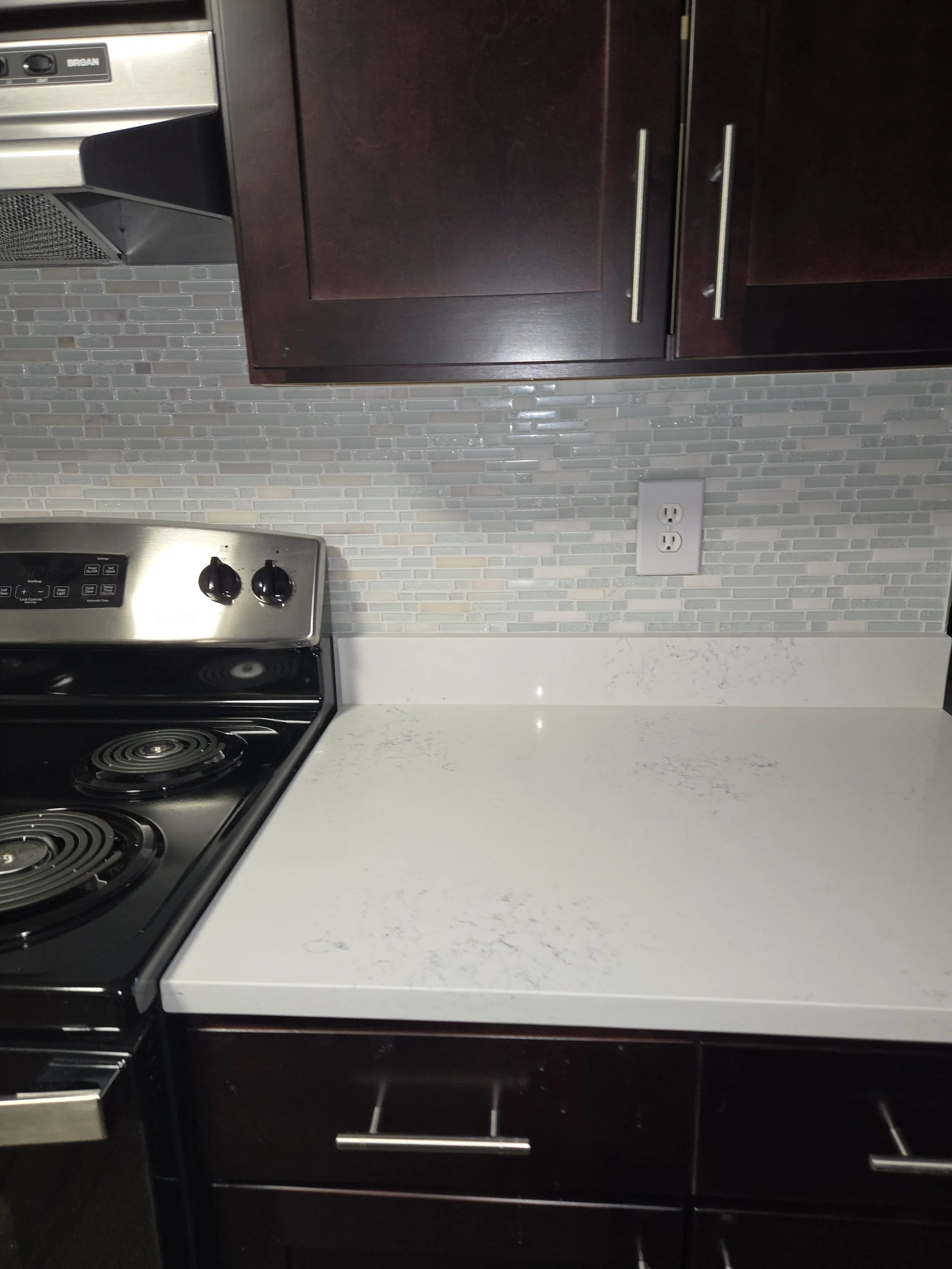A kitchen countertop with a stovetop featuring four burners, dark wood cabinets with silver handles above, a gray mosaic tile backsplash, and an electrical outlet on the wall.