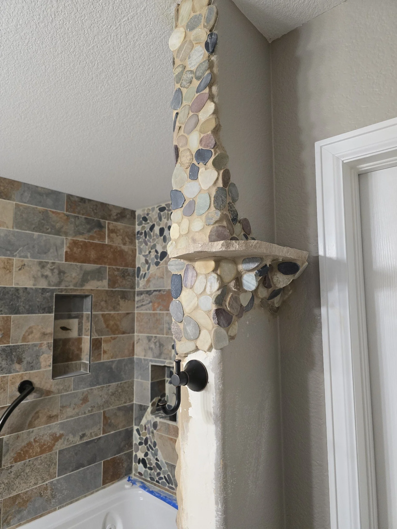A bathroom with a pebble stone accent wall, a beige countertop with a rustic edge, and a black faucet.