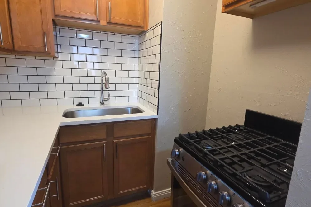 Kitchen with white countertop, wooden cabinets, white subway tile backsplash, stainless steel sink, and a black gas stove.