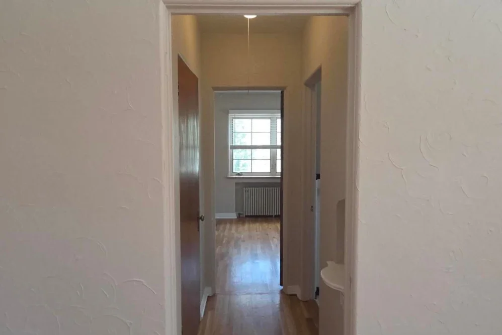 Indoor hallway view leading to a room with a window and hardwood floor.