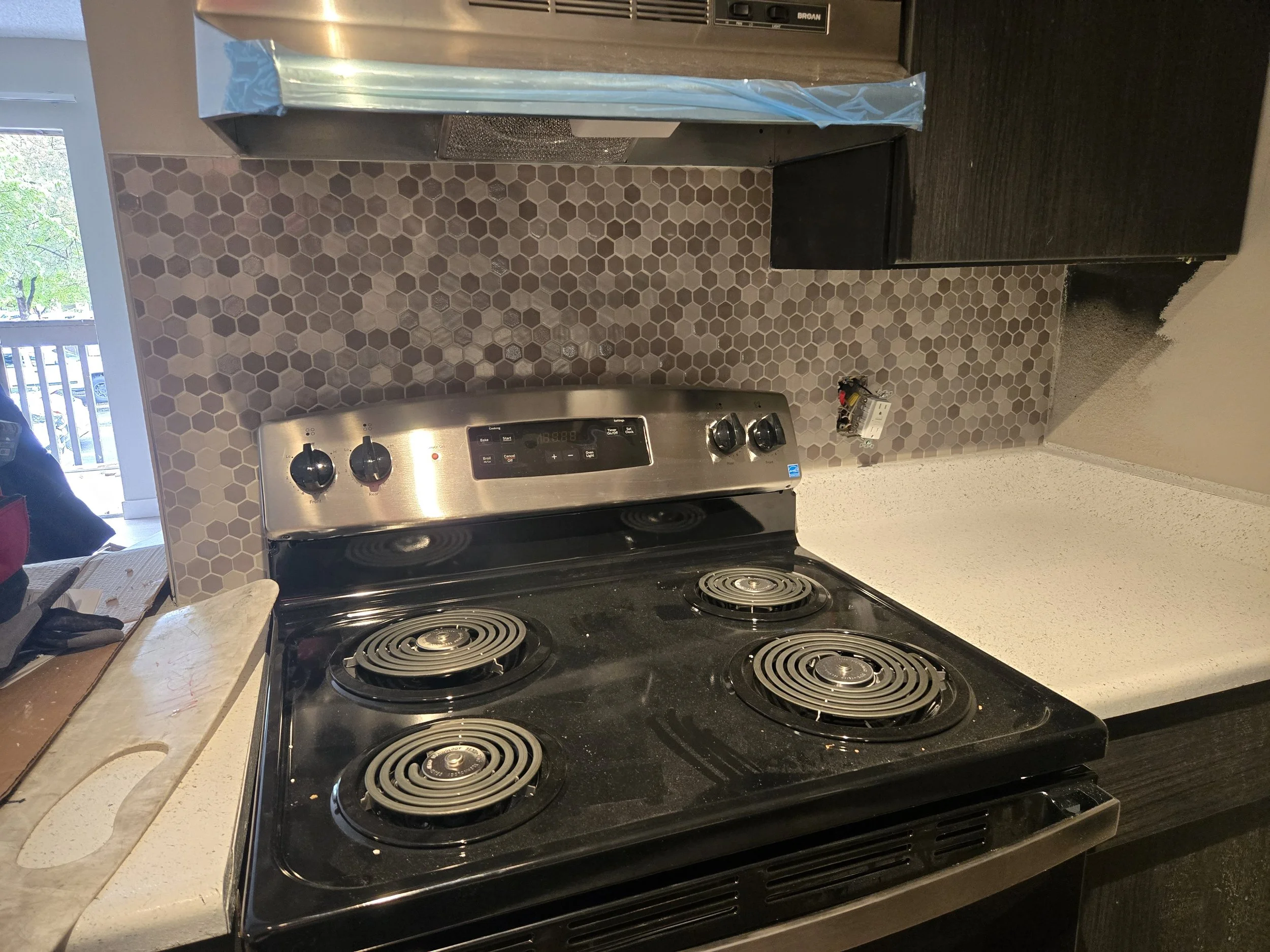A black electric stove with four coil burners, a stainless steel control panel with knobs, and a backsplash with hexagon-shaped tiles in shades of beige and brown.