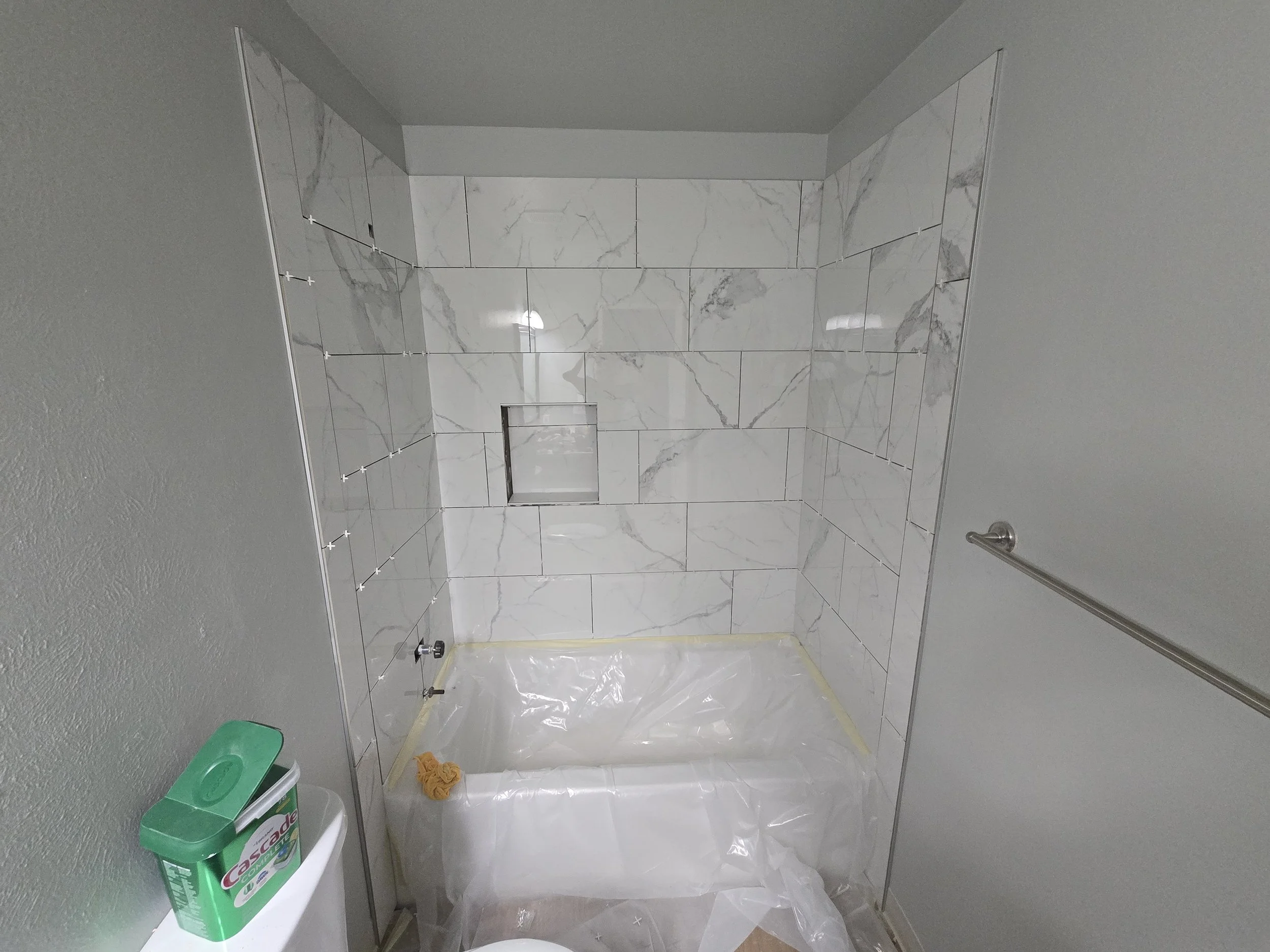Bathroom with white marble tiles on walls and floor, shower area with a niche, gray painted ceiling, a toilet paper holder, cleaning supplies, and a yellow cleaning cloth on the floor.