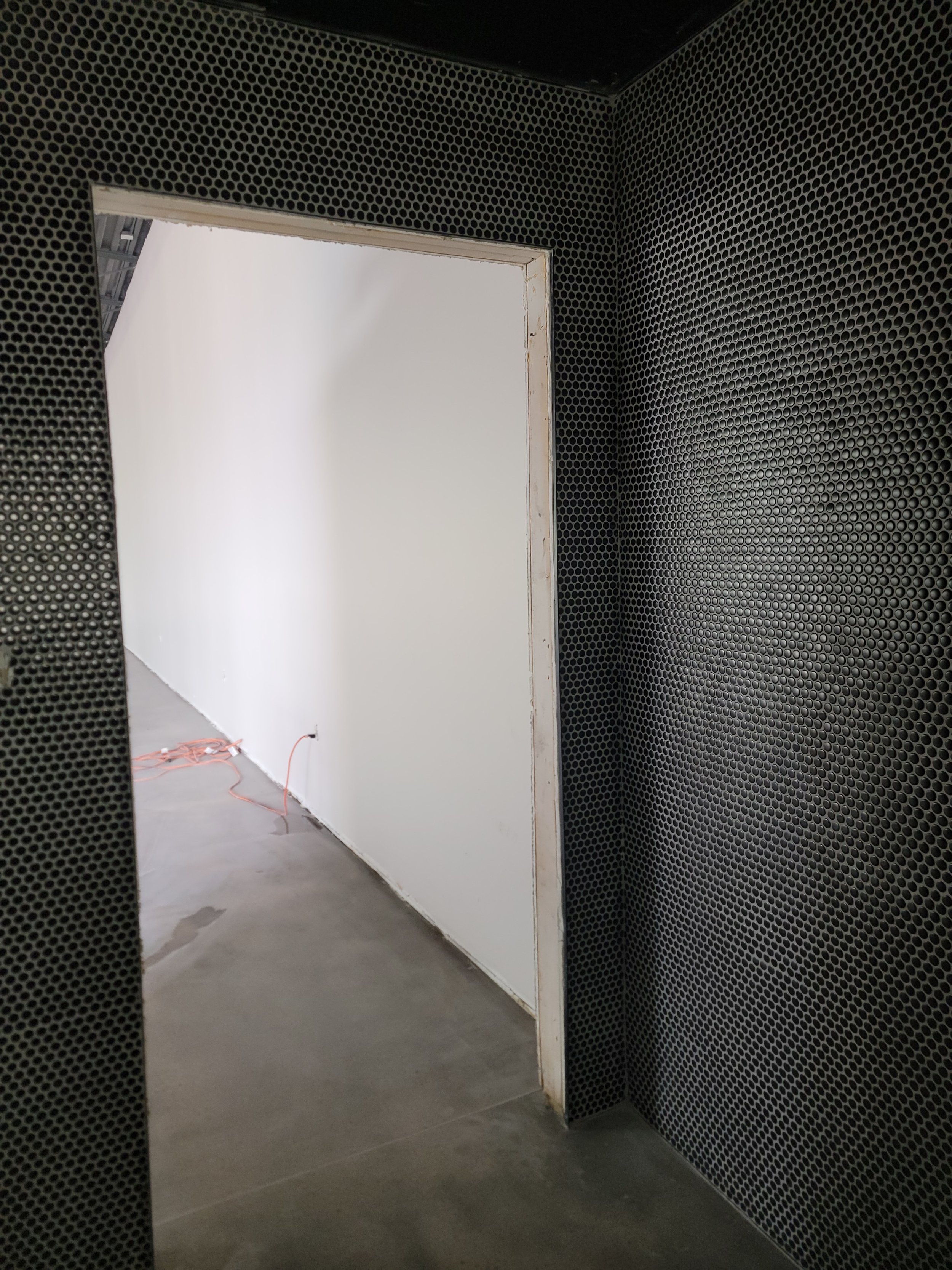 Interior of a room or hallway under construction with black soundproofing foam on the walls and electrical wiring on the floor, visible through an opening in the wall.