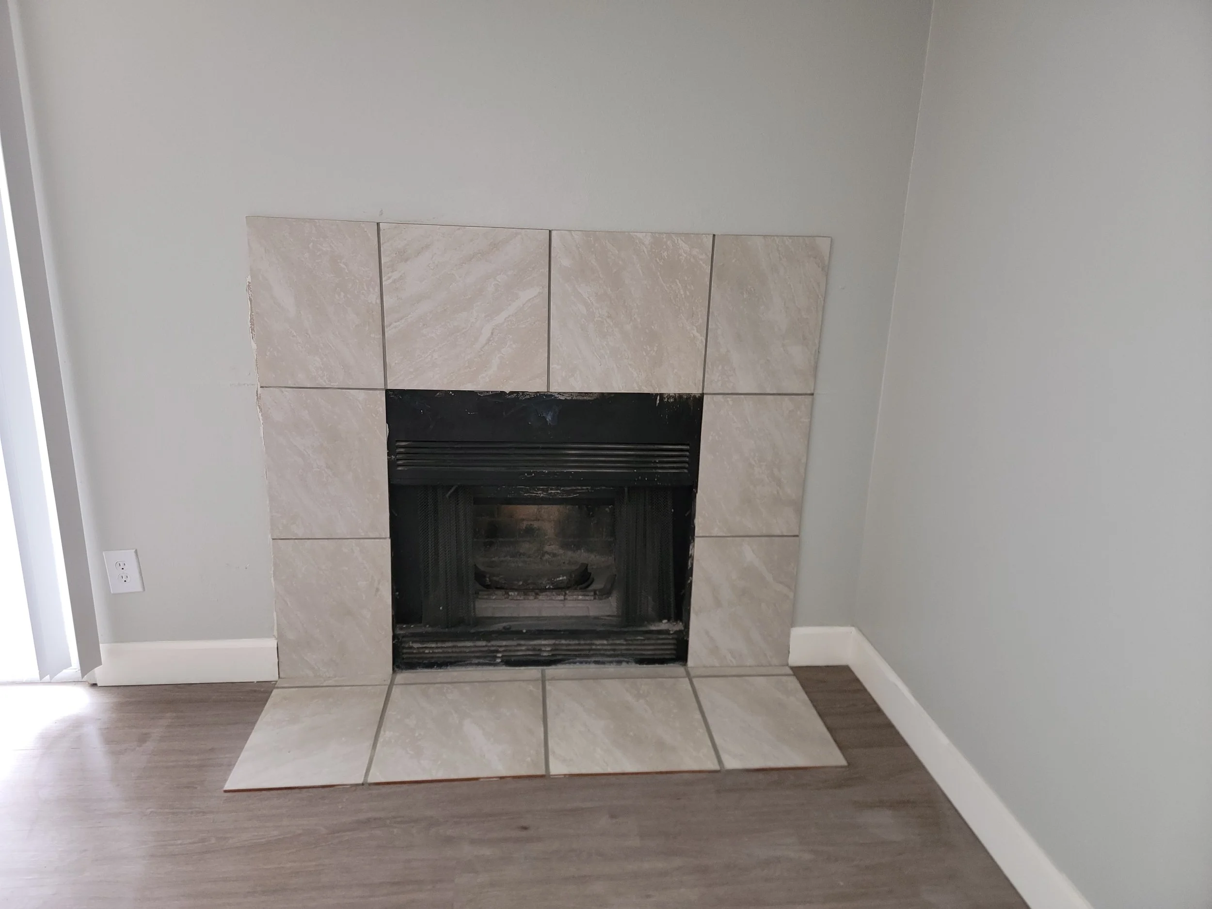 Empty fireplace with beige tiles and a black metal grate, against a light gray wall in a room with wood laminate flooring.