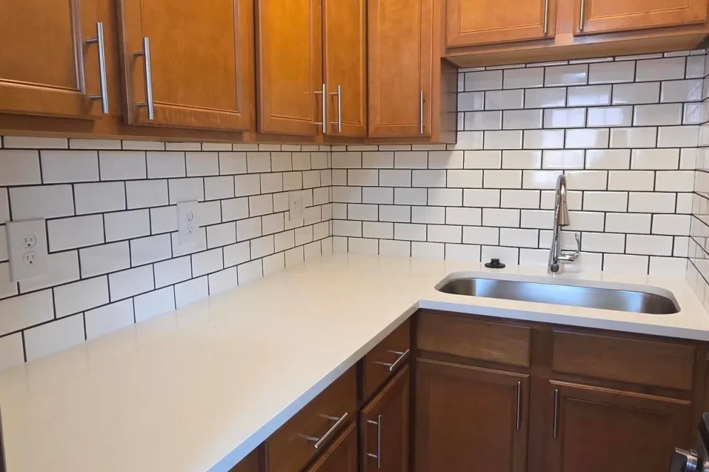 Kitchen with wooden cabinets, white countertop, and a stainless steel sink with a chrome faucet. The backsplash features white subway tiles with dark grout.