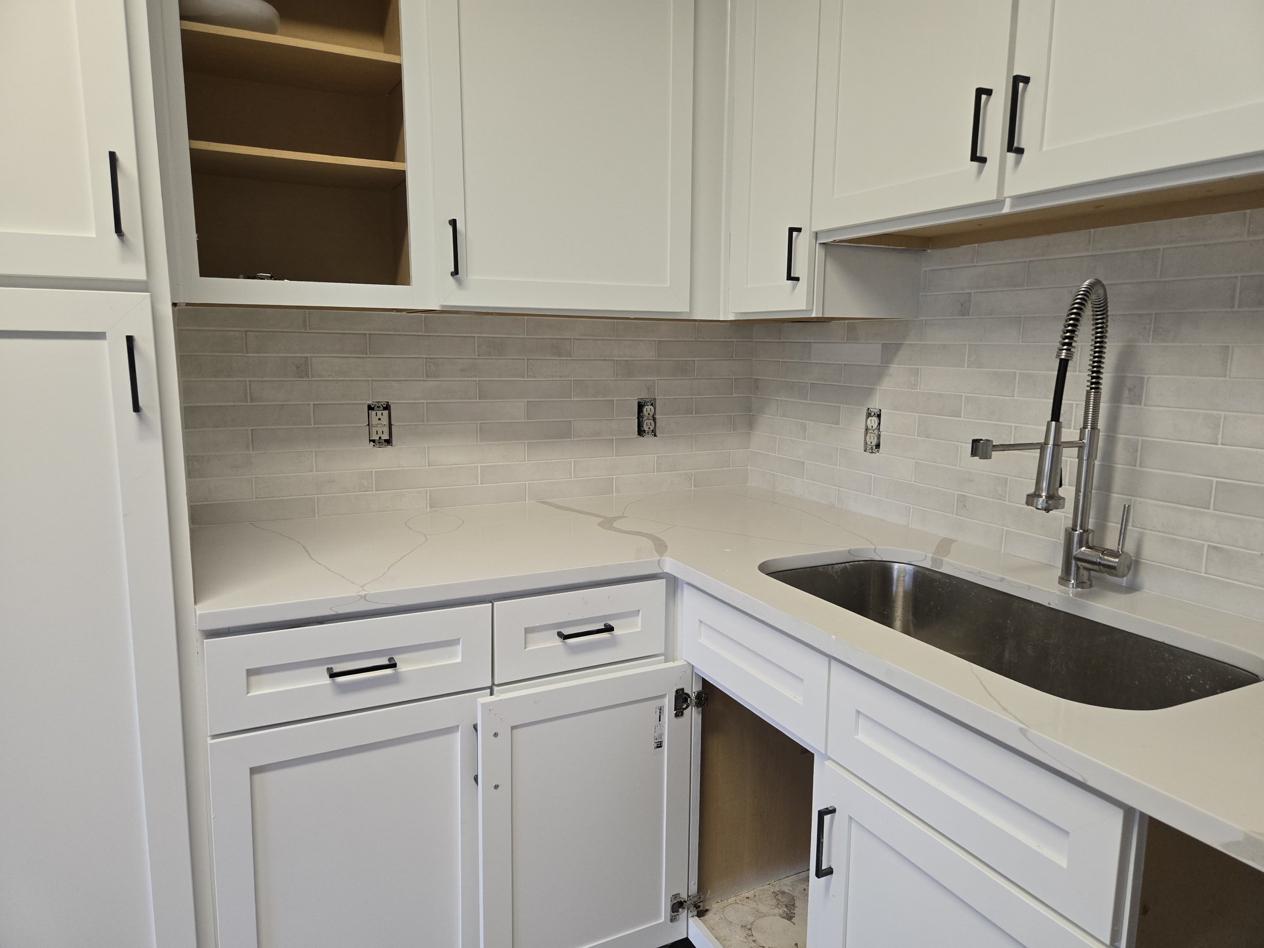 Kitchen area with white cabinets, a sink with a modern faucet, electrical outlets, and a backsplash with beige tiles. Some cabinet doors are open or missing.