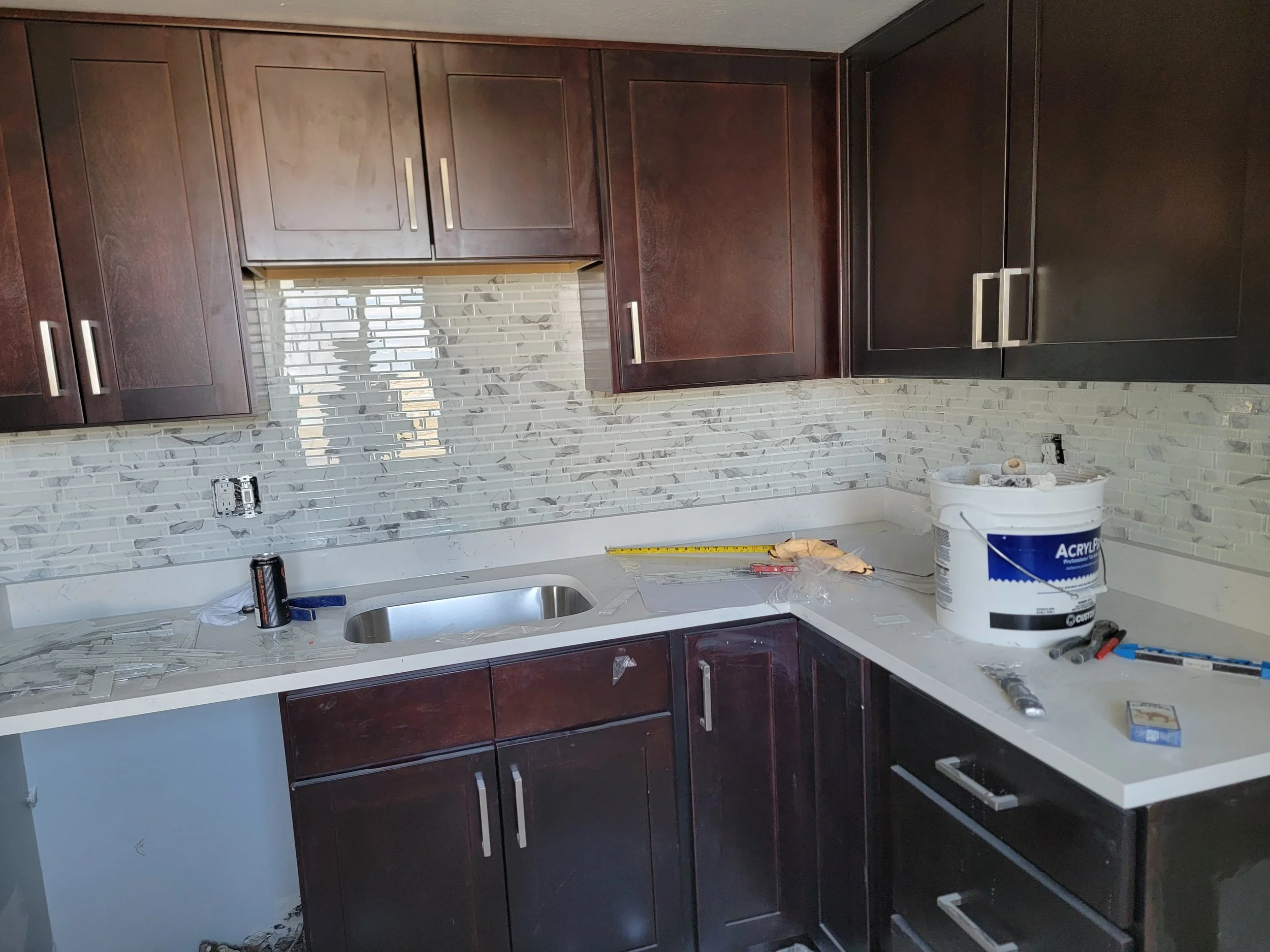 Kitchen with dark brown cabinets and a gray marble tile backsplash under construction. Tools and materials are on the white countertop, including a can of acrylic primer, a protective glove, and various small tools.