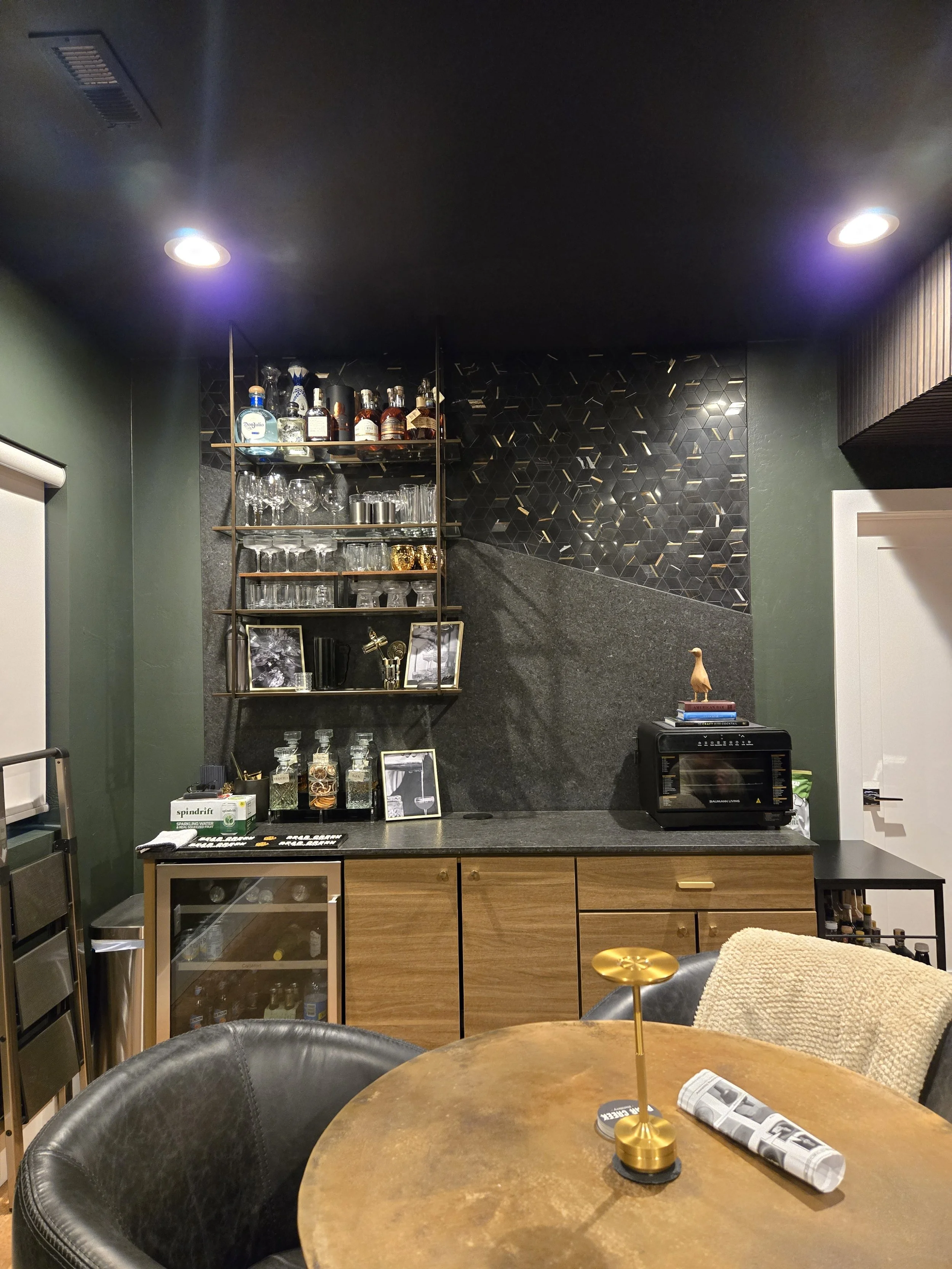A cozy bar area with wine and liquor bottles, glassware, and framed photos on a black wall, with a black countertop. In the foreground, a round wooden table has a gold decorative object, a stress ball, and a rolled-up newspaper. There is a black leather chair with a beige blanket draped over it and part of a gray chair visible.
