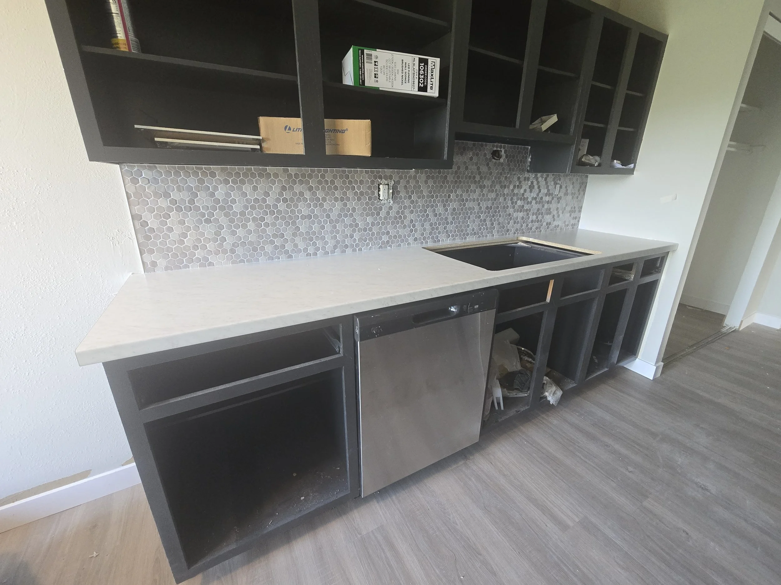 Unfinished kitchen with black cabinets, a hexagonal tile backsplash, and a white countertop. The lower cabinets and drawer handles are missing, and there are some tools and boxes on the open upper shelves. The flooring is gray wood.