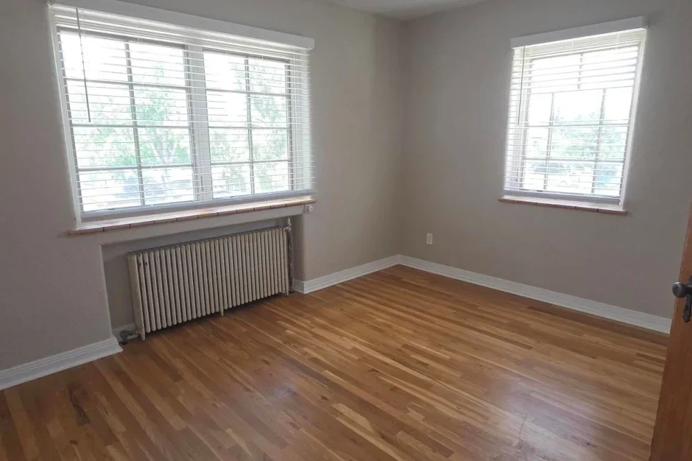 Empty room with hardwood floors, two large windows with blinds, and a radiator beneath one window.