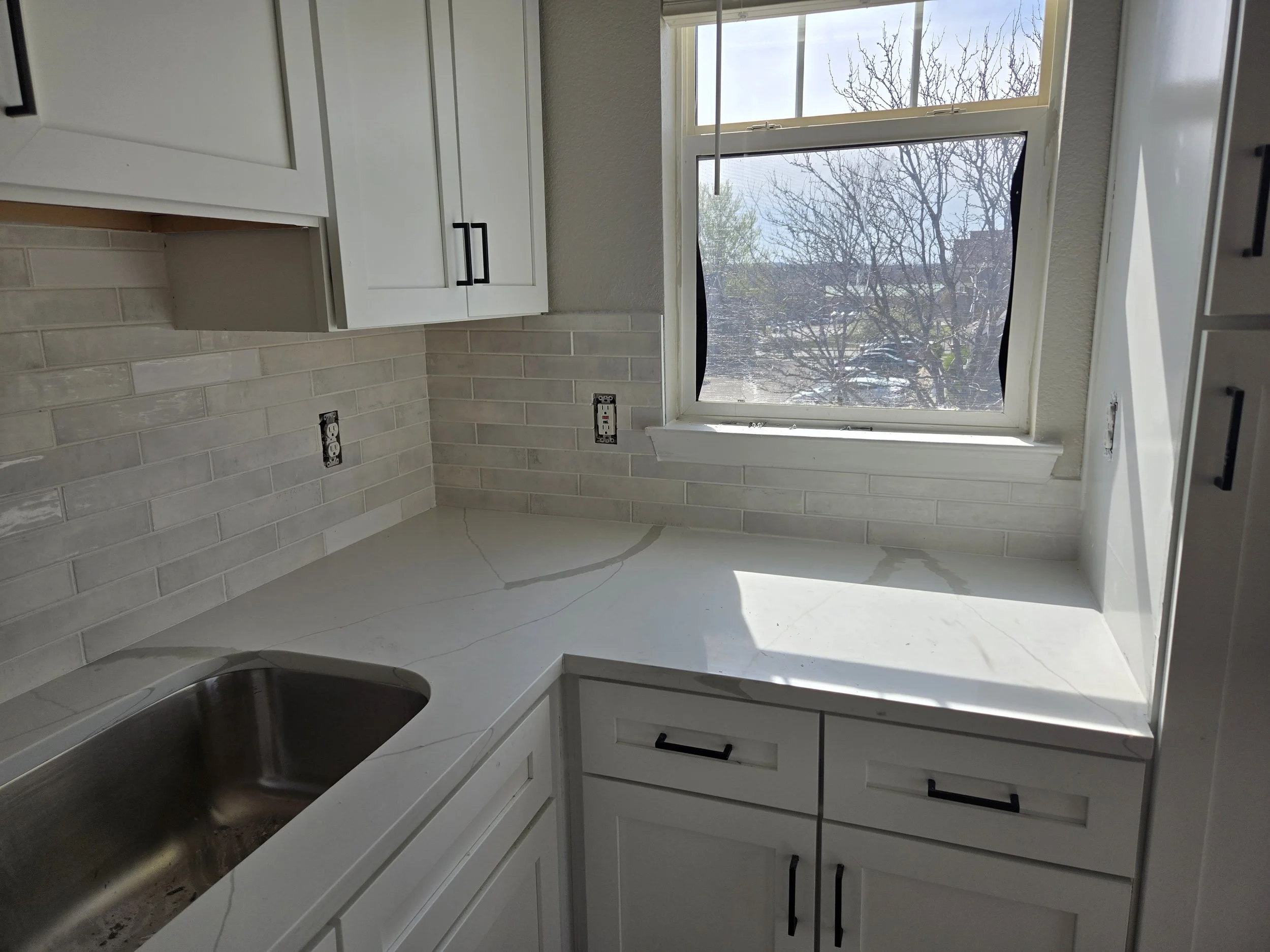 Kitchen with white cabinets and a large window, showing a view of leafless trees outside. The kitchen has a gray countertop with a stainless steel sink, and the backsplash is made of beige subway tiles. There are electrical outlets on the wall below 